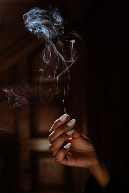 Hand holding a lit incense stick with smoke curling upward in a dimly lit room.
