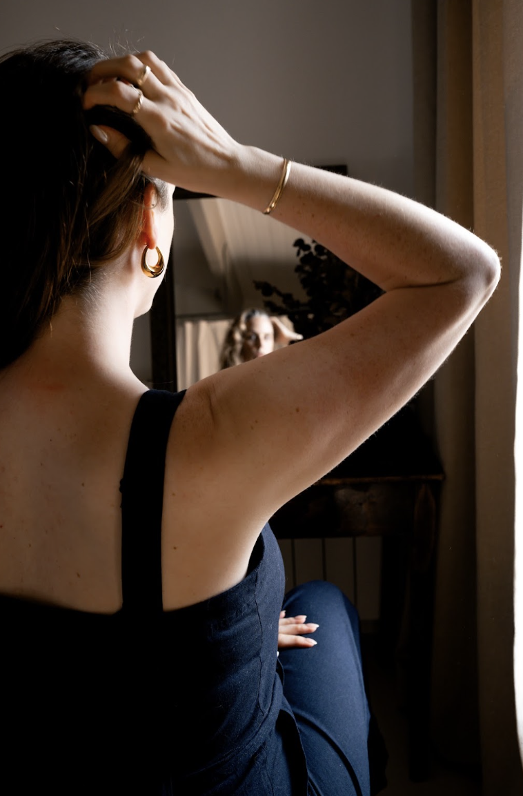 A woman sits by a window, looking at her reflection in the mirror, with her right hand on her head. She wears a black top, a gold bracelet, and hoop earrings, with light coming through the window casting a glow on her hair and skin.