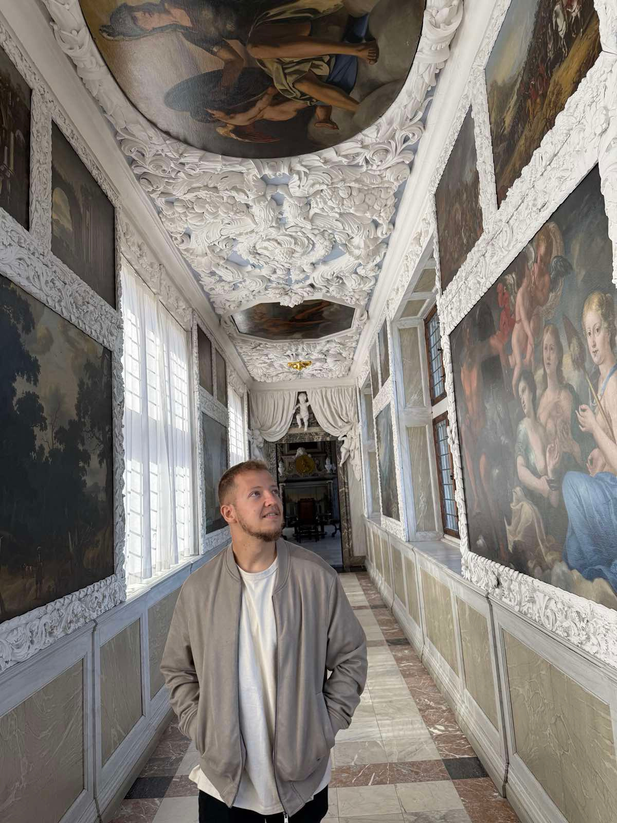 A young man with short hair and a beard looking up inside a decorated art gallery or museum, surrounded by large framed paintings on ornate white walls with intricate molding and a ceiling with elaborate white stucco designs and paintings.