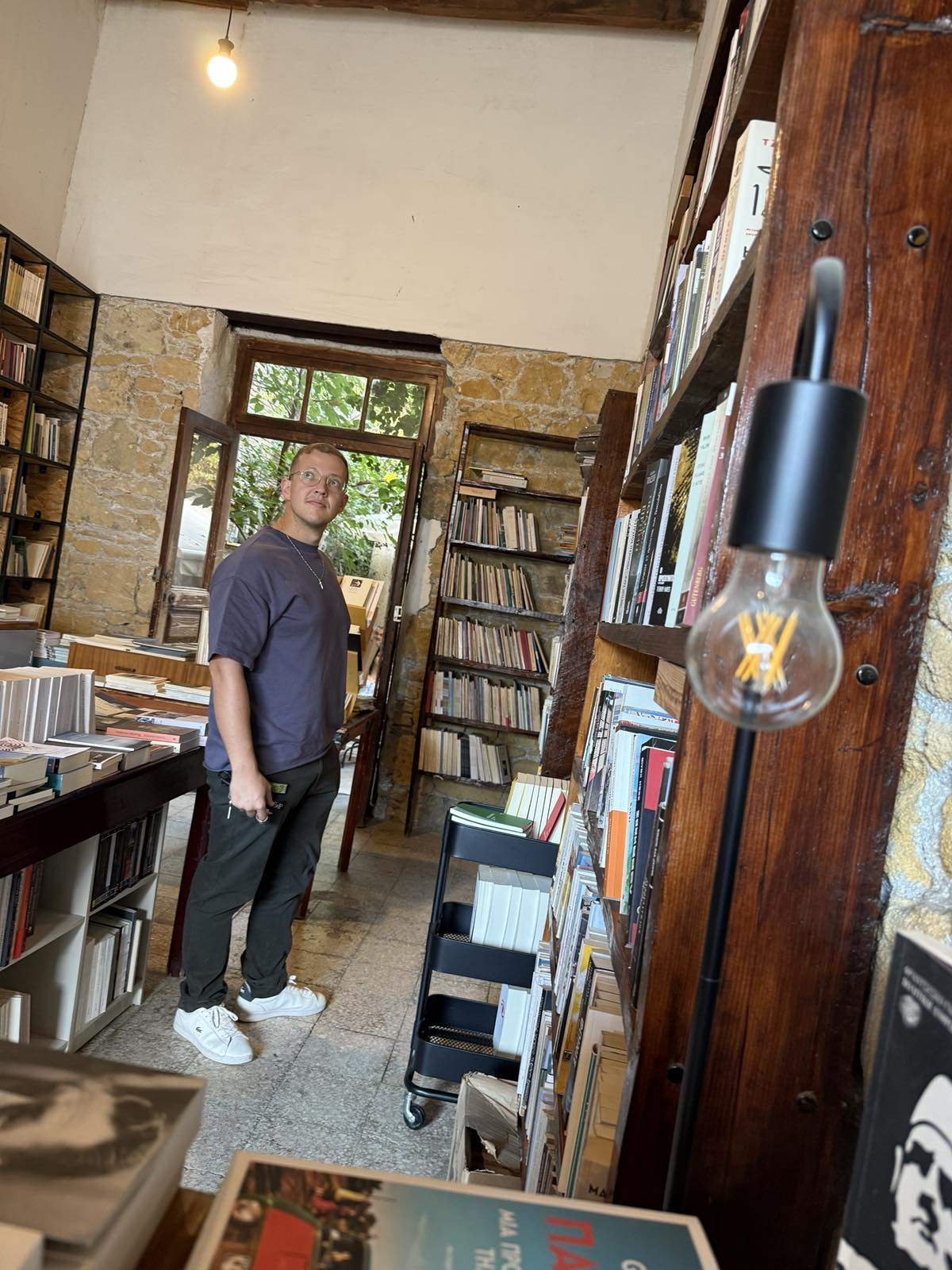 A man in a bookstore surrounded by shelves of books, looking at the camera. The store has exposed brick walls, a window open to green foliage, and warm lighting.
