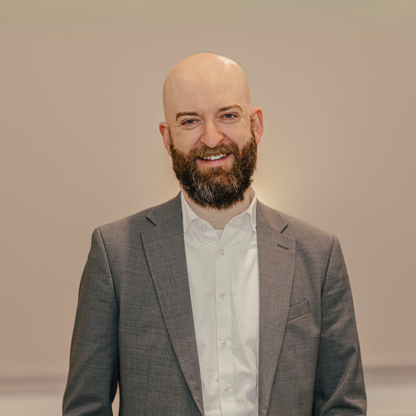 Ein Mann trägt einen grauen Anzug, eine blaue Hemd und einen Hut, steht im Freien auf einer sonnigen Wiese mit grünen Bäumen im Hintergrund.