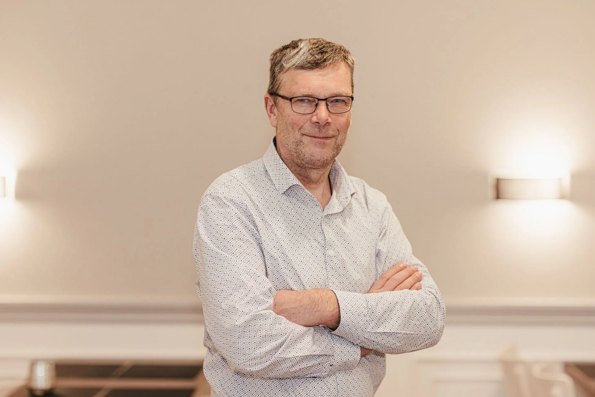 Ein Mann in einem grauen Hoodie steht lachend im Freien vor einer Wiese mit Hochspannungsleitungen und einer Industrieanlage im Hintergrund.