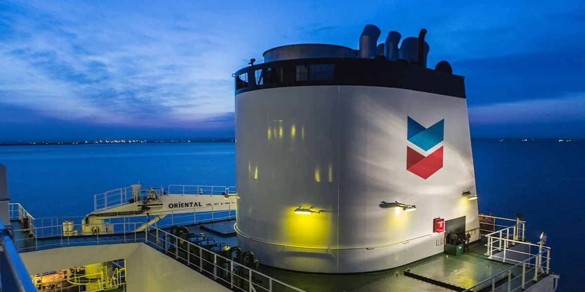 The exhaust stack of a Chevron ship in a large coastal port area at dusk. Lights illuminate the Chevron logo.