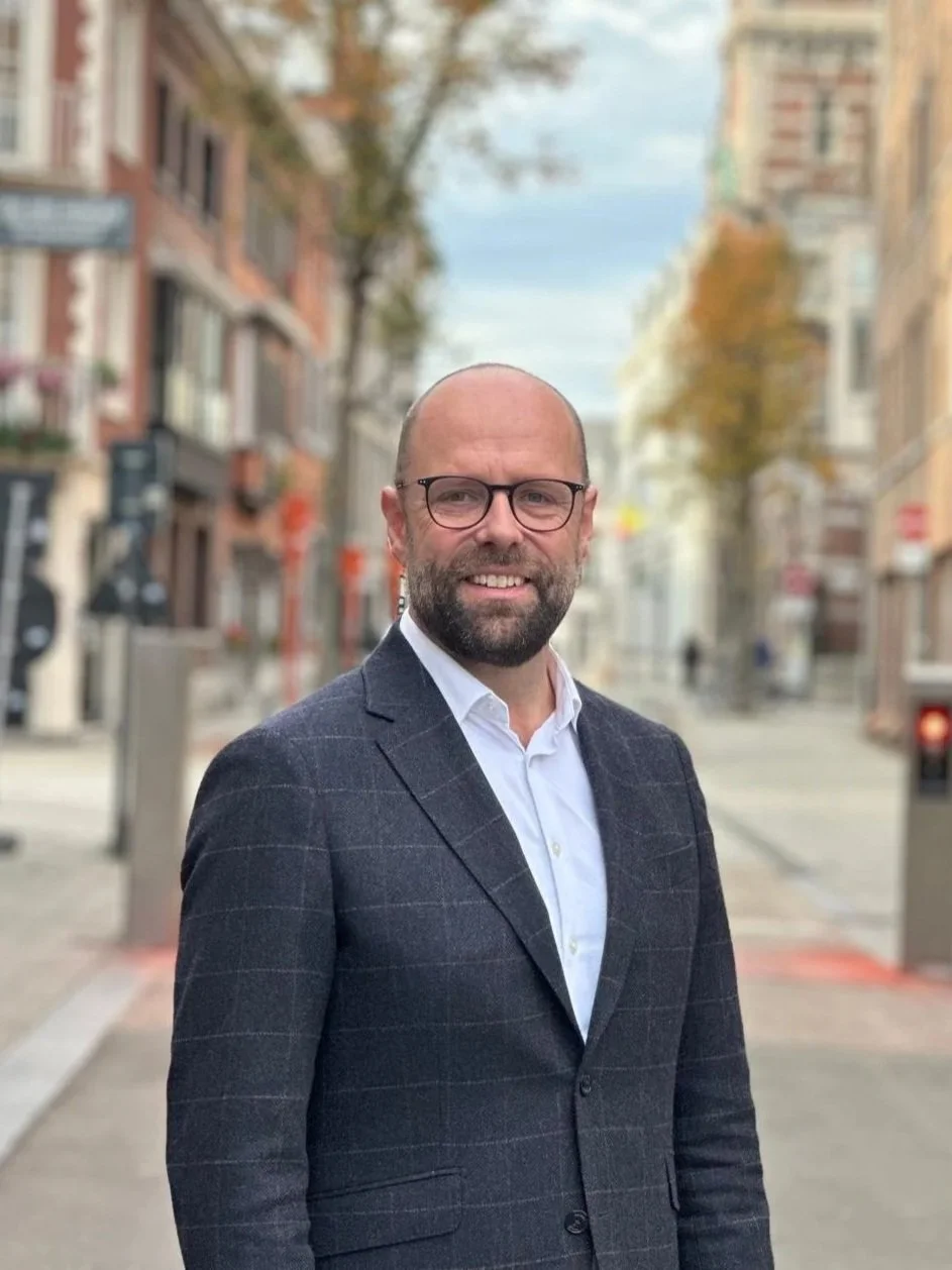 Tom Thijsen, man met bril, baard en korte haar, lachend, in een stad met oude gebouwen en bomen in de achtergrond.
