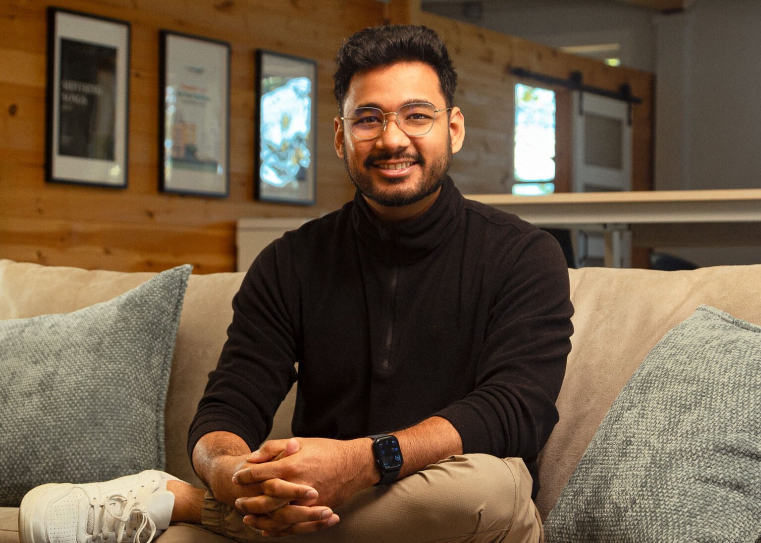 A young man with glasses and a beard sitting on a beige couch with gray pillows in a cozy indoor space with wooden walls, smiling at the camera.
