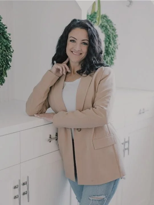 A woman with dark, curly hair; smiling, standing indoors by a white cabinet with green plants in the background.