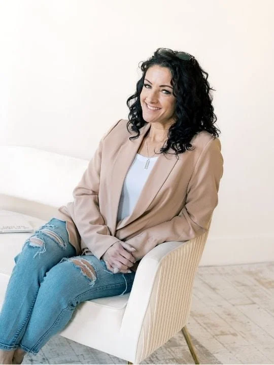 A woman with curly dark hair sitting on a cream-colored armchair, smiling, wearing a tan blazer, white shirt, and ripped jeans, in a bright room.