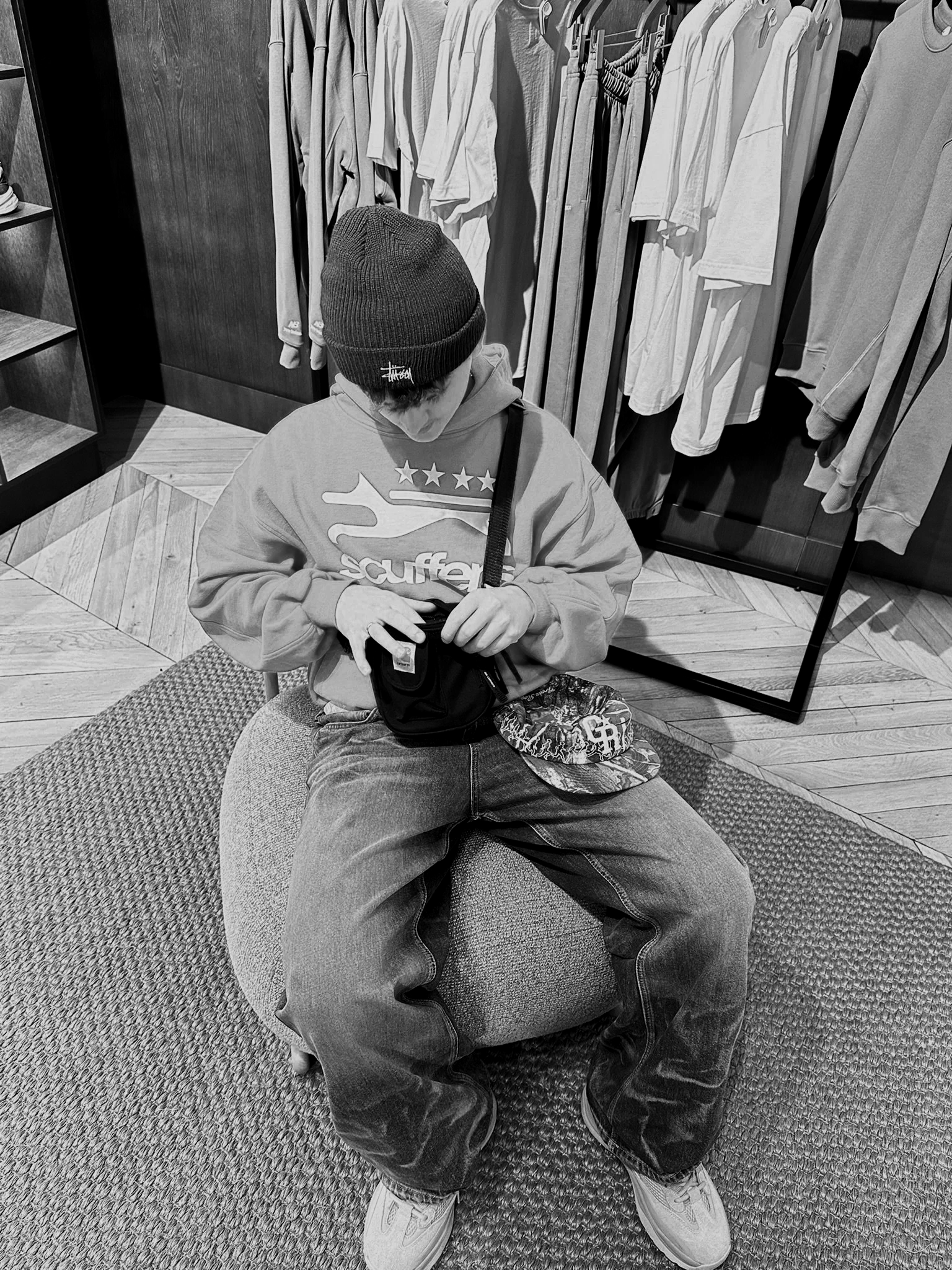 A young person sitting on an oval-shaped cushioned seat in a clothing store, wearing a beanie, hoodie, jeans, and sneakers, with a small bag and a patterned cap on their lap. They are looking into the bag, with clothing on a rack behind them.