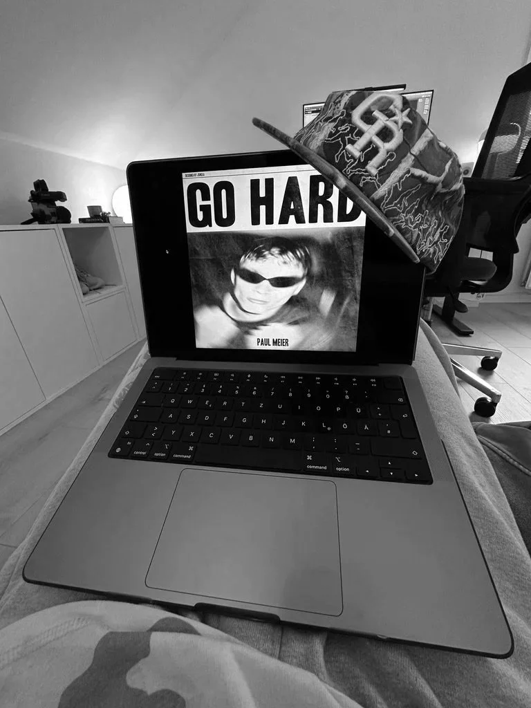 A laptop showing a black and white album cover with the text 'GO HARD' and a person's face wearing sunglasses. A baseball cap with a lightning bolt design is hanging on the laptop. In the background, there is a workspace with an office chair, a computer monitor, and a cabinet.