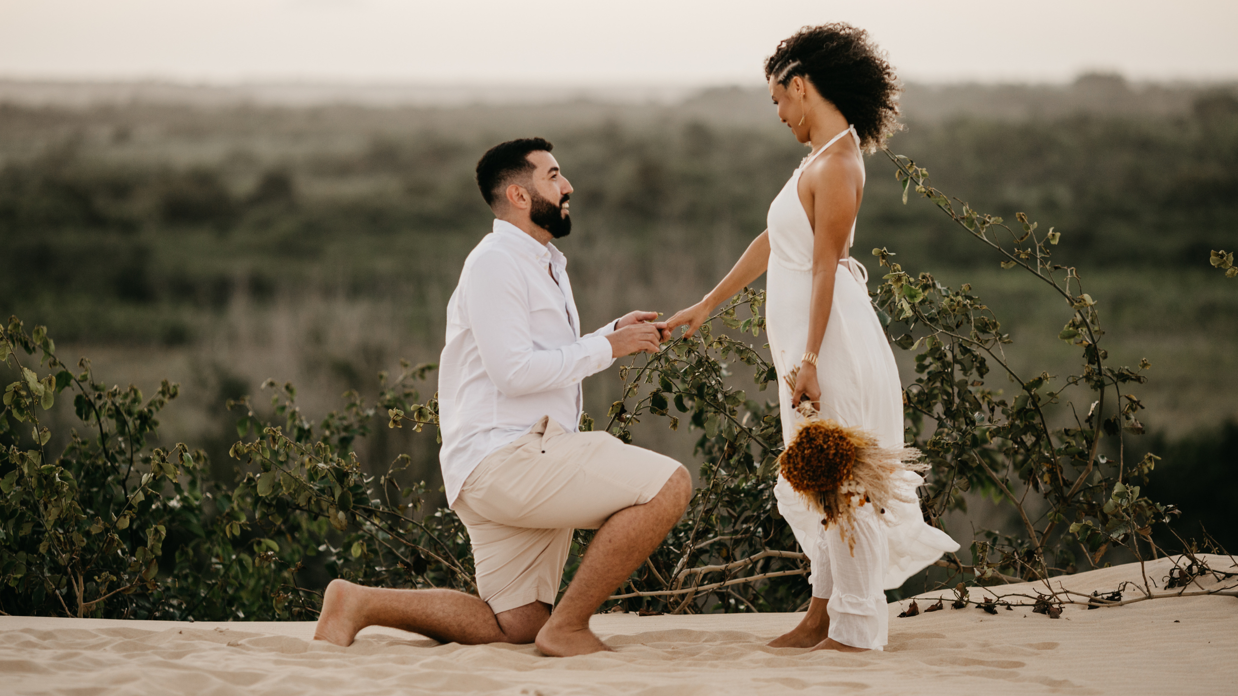 A man proposing marriage to a woman on a sandy beach with greenery in the background during sunset, with the man kneeling and the woman standing.