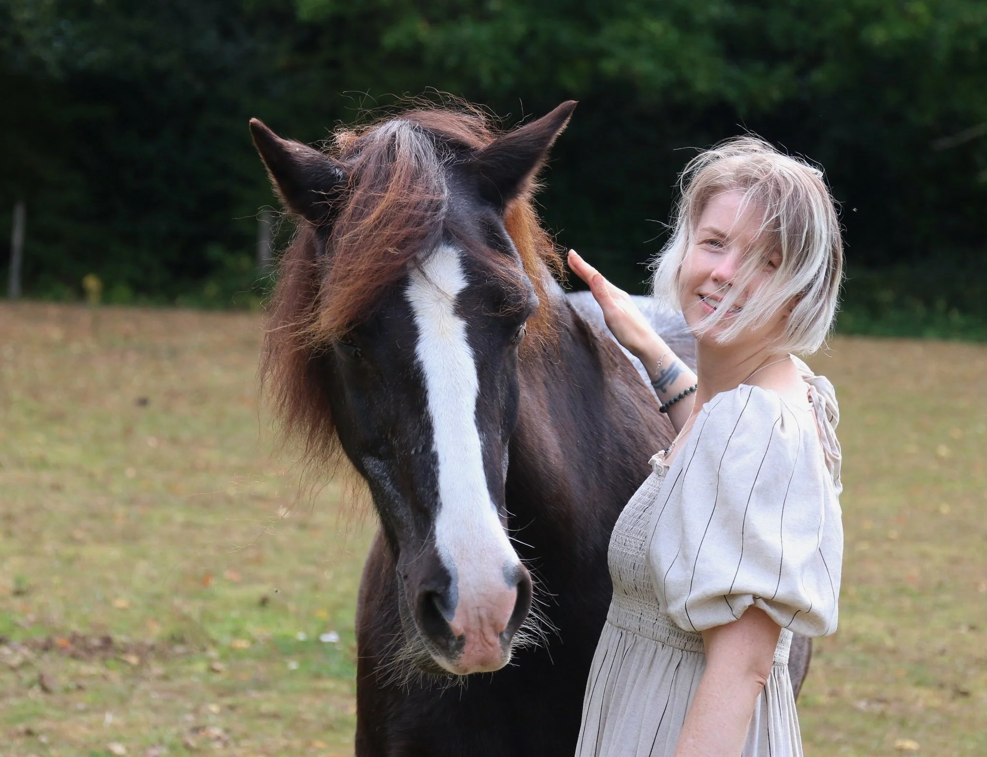 A smiling woman with short blonde hair patting a dark brown horse in a field.