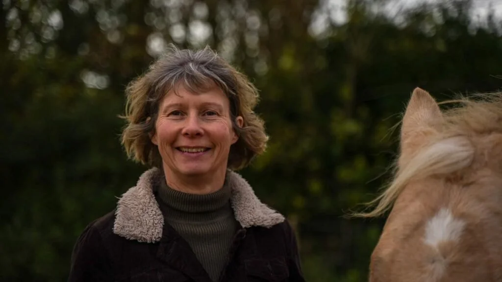 A woman with short hair and a fringe blowing in the wind standing next to a horse and smiling.
