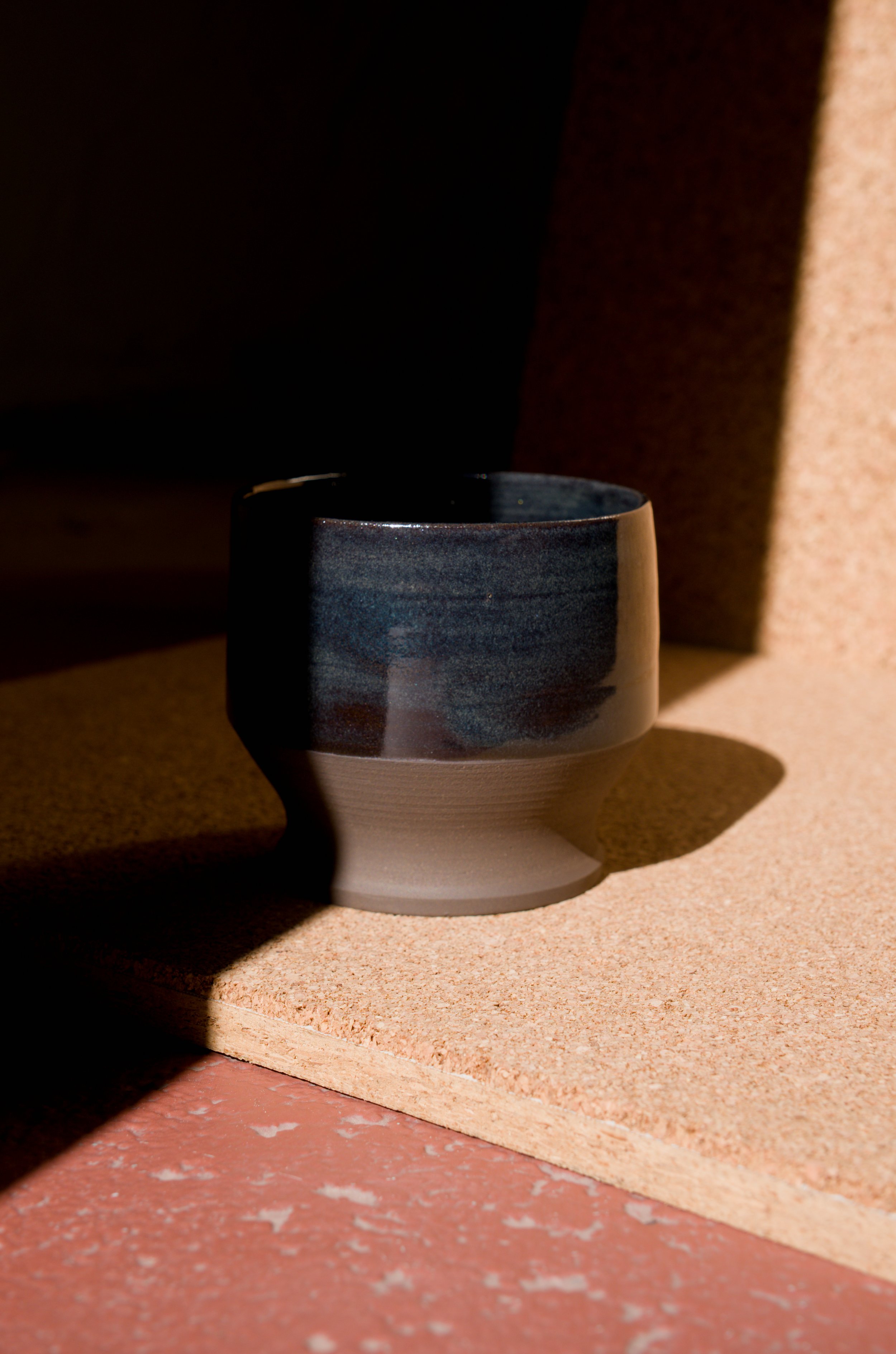 Une tasse bleue en céramique sur une surface en liège, avec un fond sombre et une lumière du soleil créant un contraste marqué.