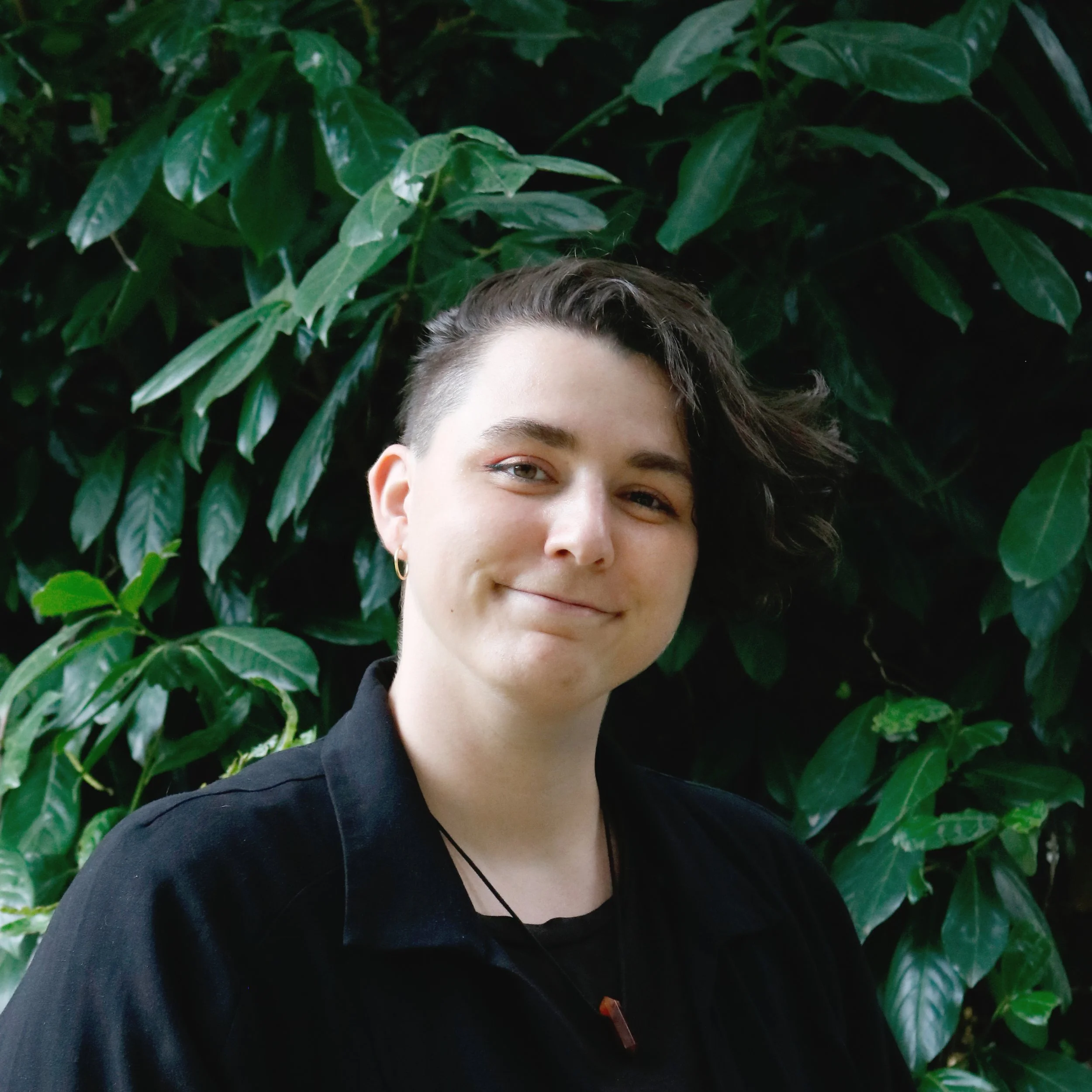 Un portrait partiel d'une personne avec des cheveux courts bouclés, des sourcils épais et un sourire subtil, posant devant un mur de feuillage vert dense.