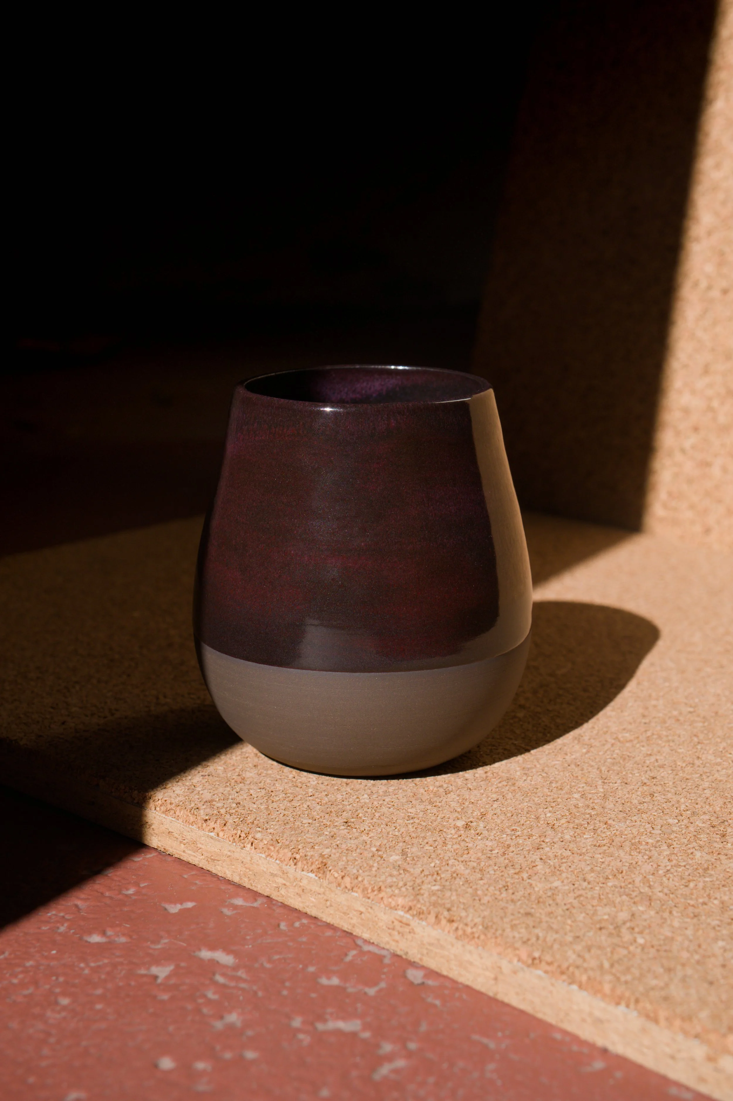 Un verre à bière en céramique à deux tons, avec une partie supérieure violette et une partie inférieure blanche, placé sur un support en bois ensoleillé.