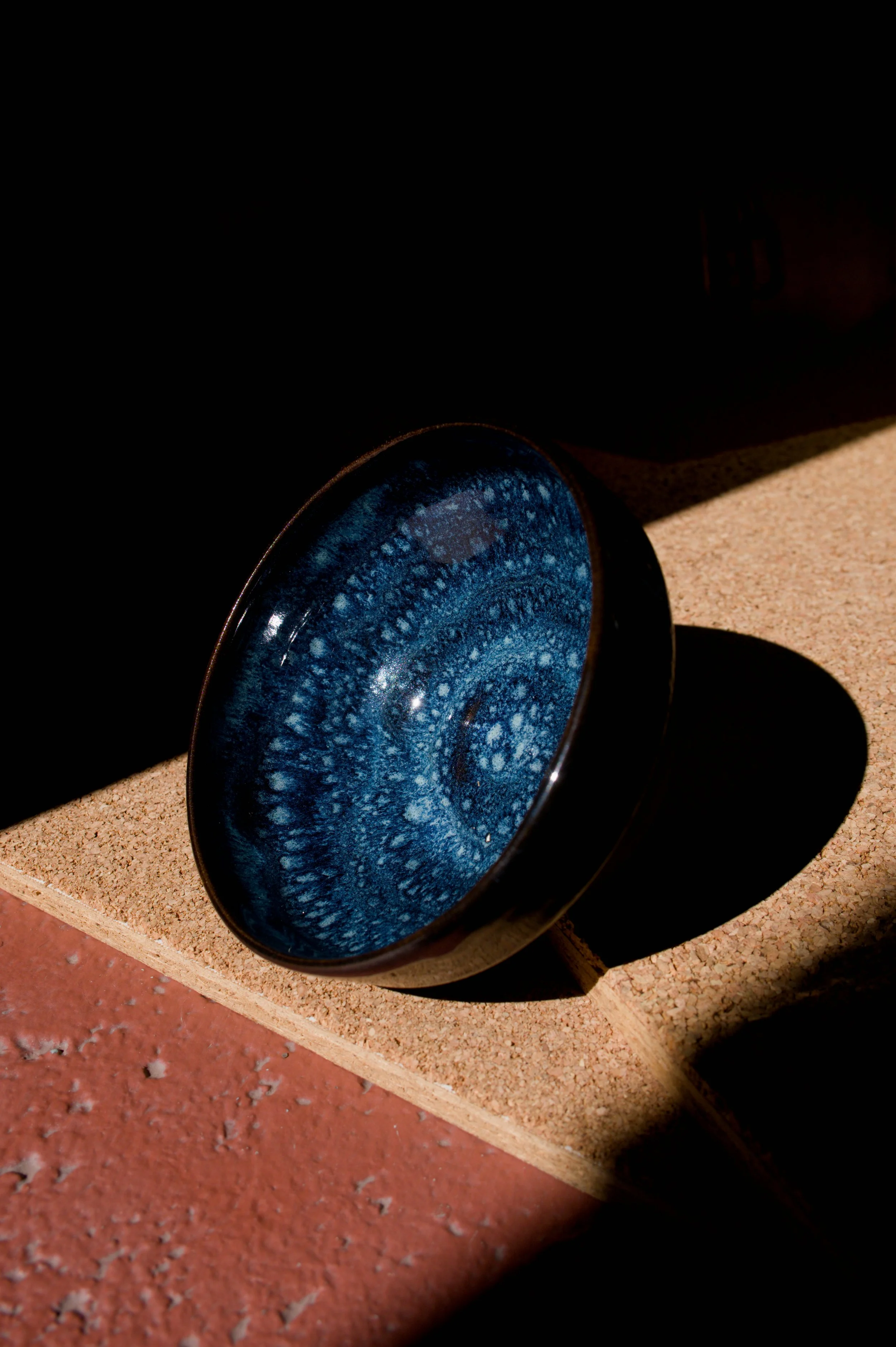 Un bol en céramique avec un intérieur bleu marbré, posée sur une surface en liège, avec un fond sombre et une ombre visible.