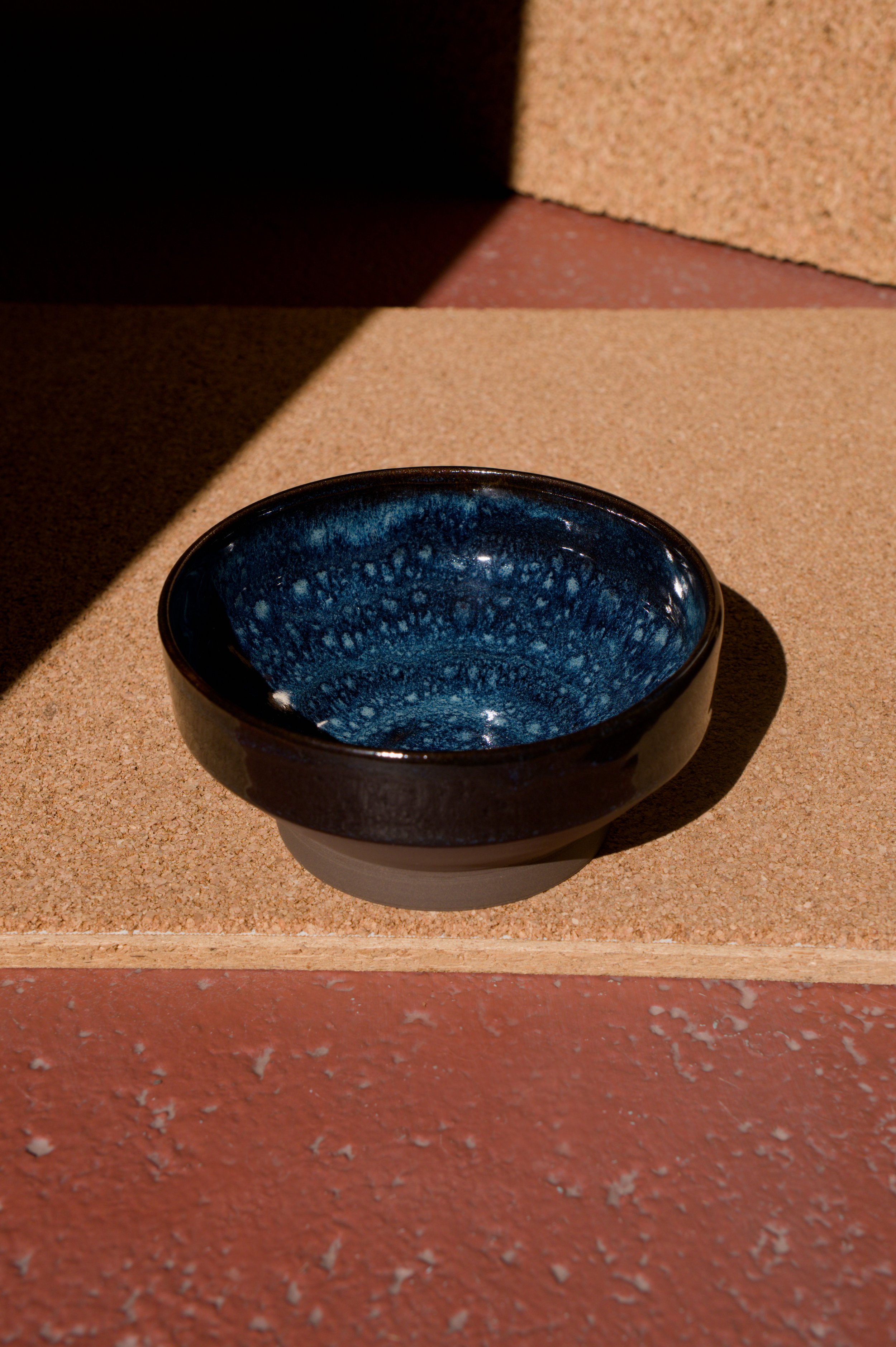 Un bol en céramique noir avec une interior bleu marine, placé sur une surface en pierre rouge et beige.