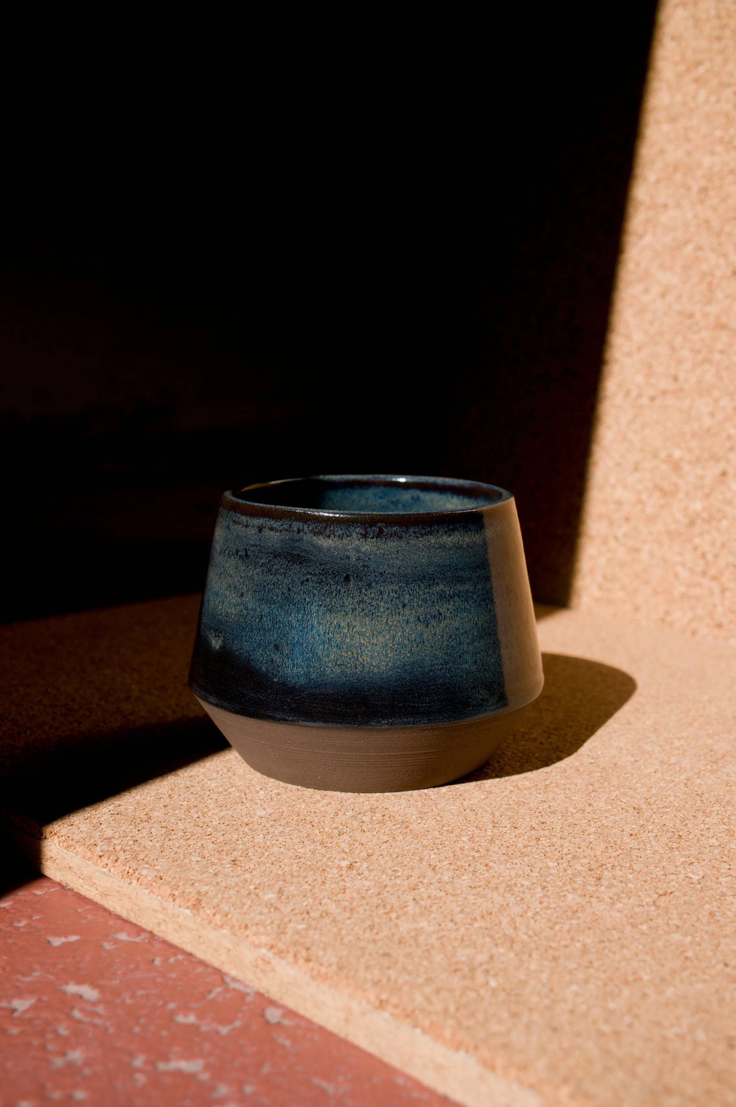 Une tasse en céramique bleue avec un dégradé de jaune et de noir, placé sur une surface en liège, avec un fond ombragé créant un contraste entre la lumière et l'ombre.