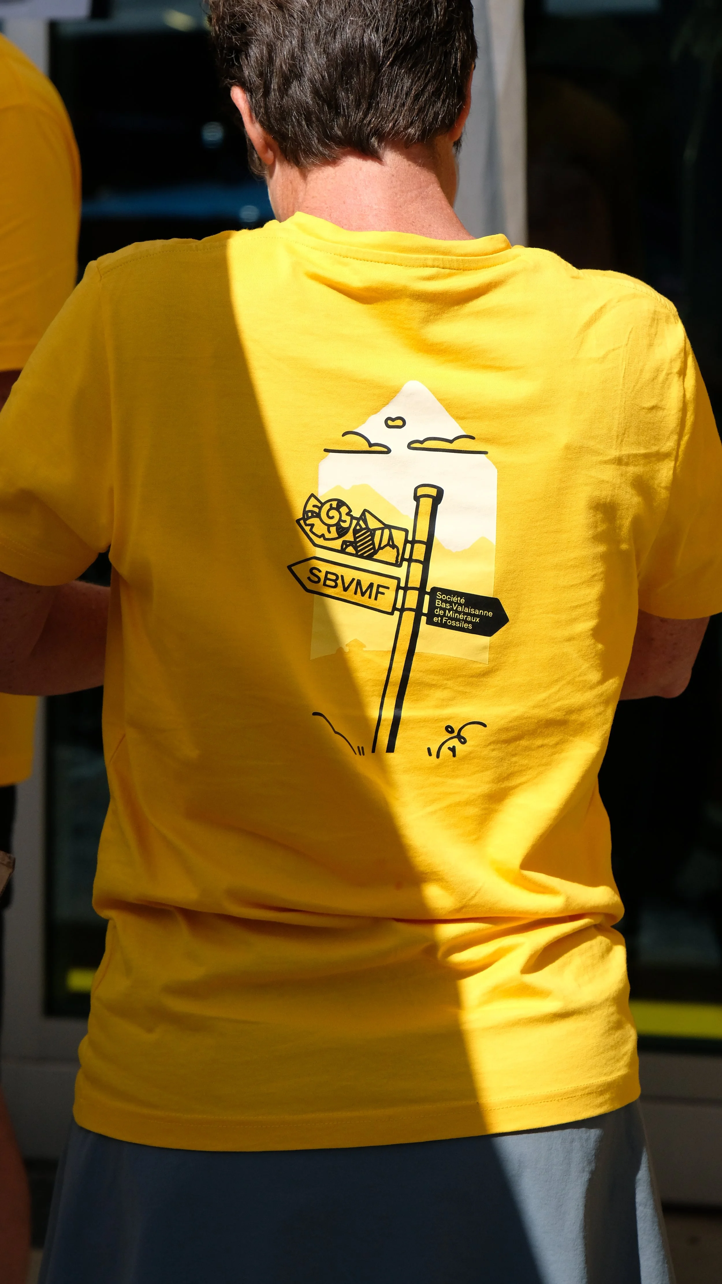 Une personne porte un t-shirt jaune avec un dessin illustrant une montagne, un panneau directionnel marqué SBVMF, et une usine de minéraux et fossiles.