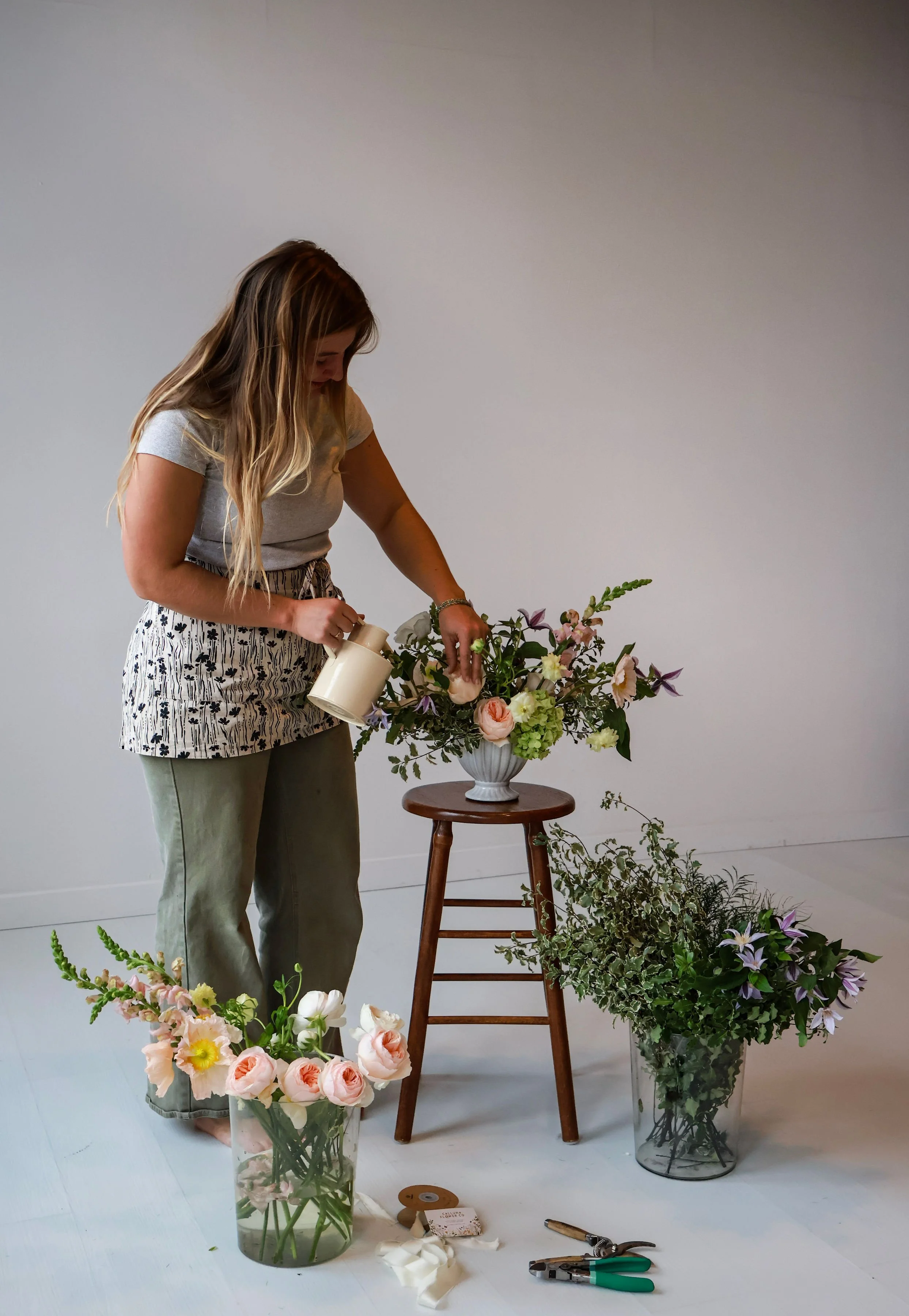 A woman arranging flowers in a white vase, surrounded by additional floral arrangements and tools on the floor, in a minimal indoor setting.
