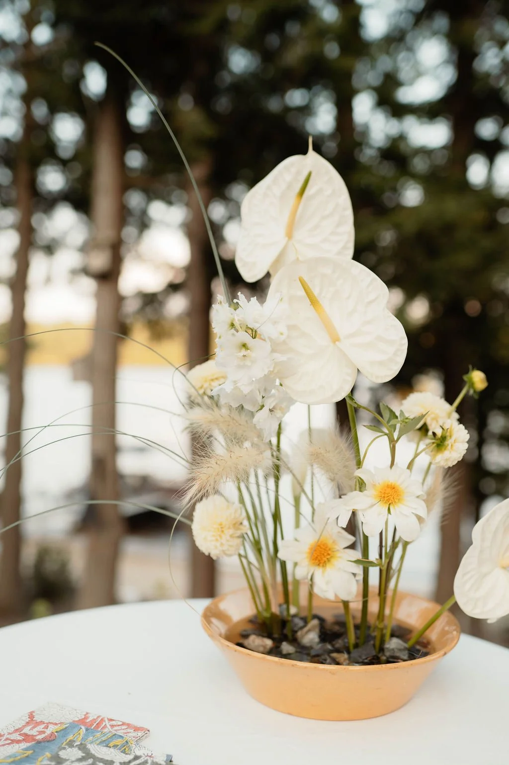 A flower arrangement in an orange ceramic bowl containing white and cream flowers, with some having yellow centers, and decorative grasses with fluffy beige tips, set against a blurred outdoor background.