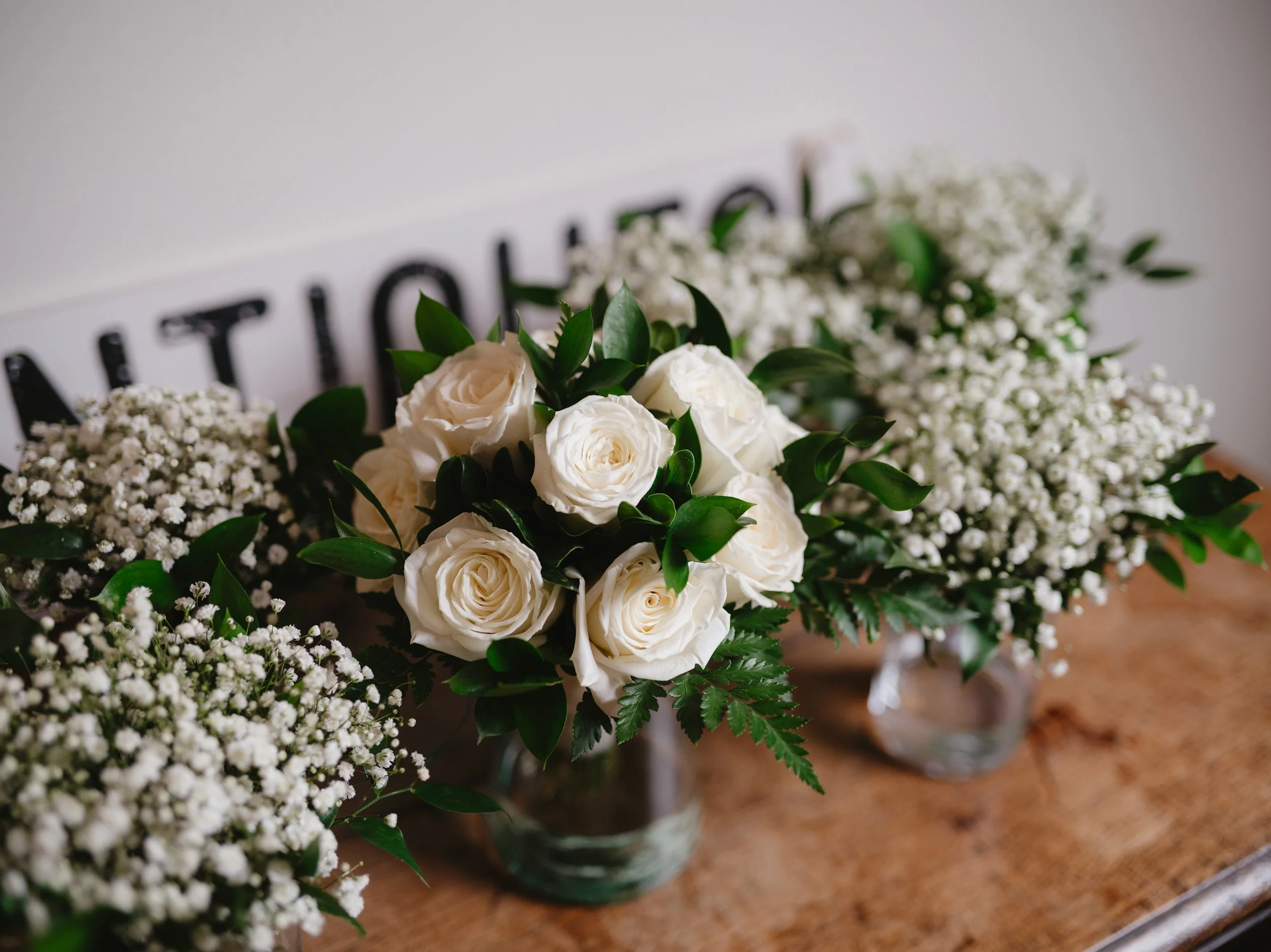 White roses and baby's breath flower arrangements in glass vases on a wooden table.