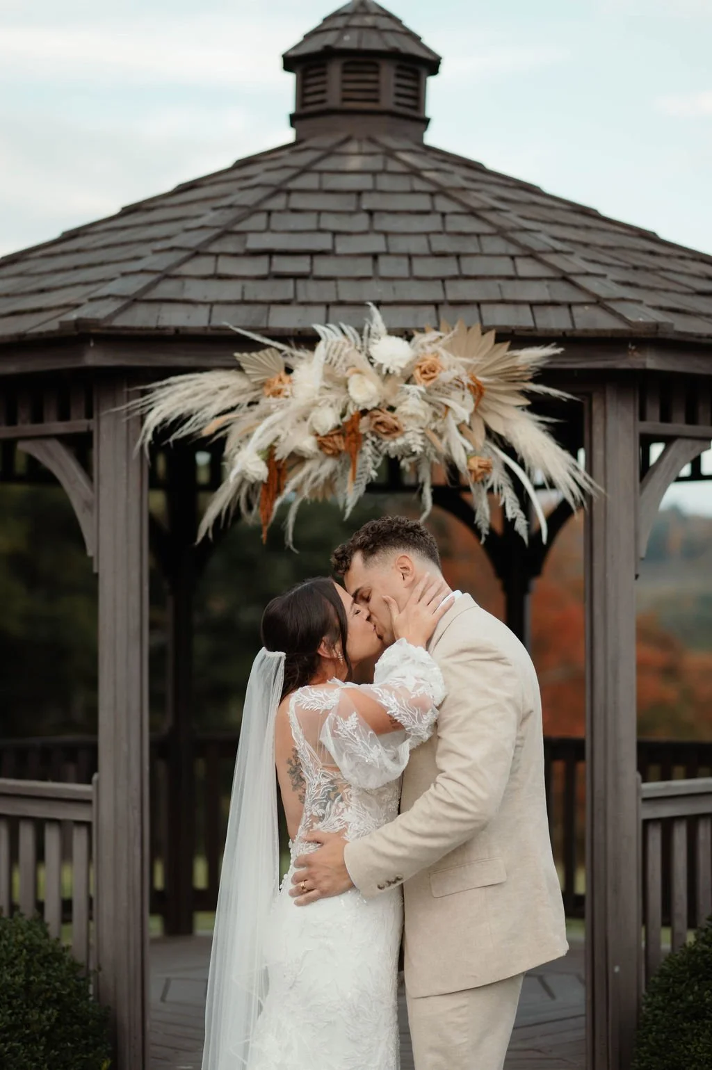 A bride and groom kissing under a gazebo decorated with a large floral arrangement at their wedding.