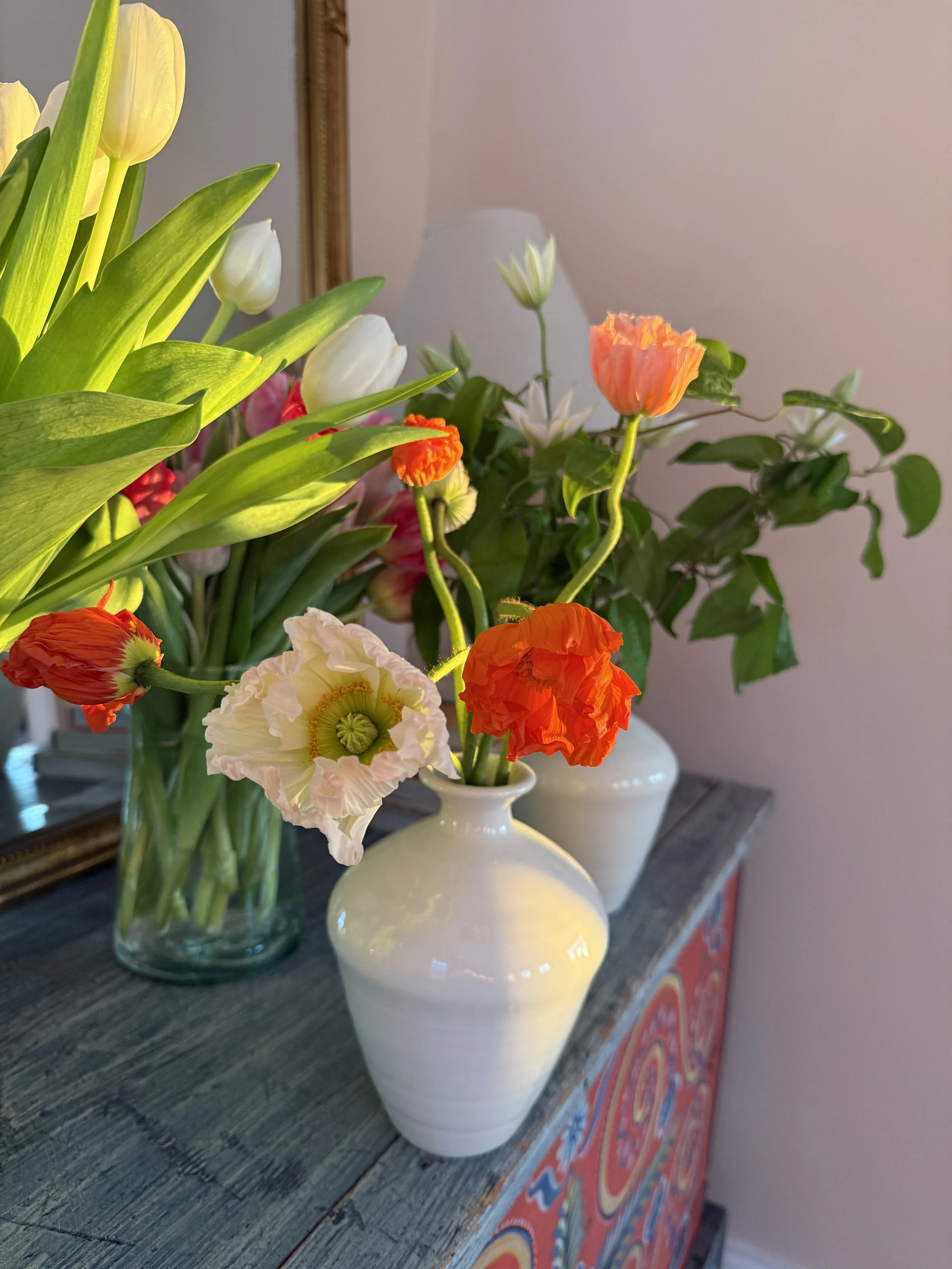 A close-up of a floral arrangement in white vases on a wooden table, featuring white, yellow, orange, and pink tulips and other flowers, with a mirror and a pink wall in the background.