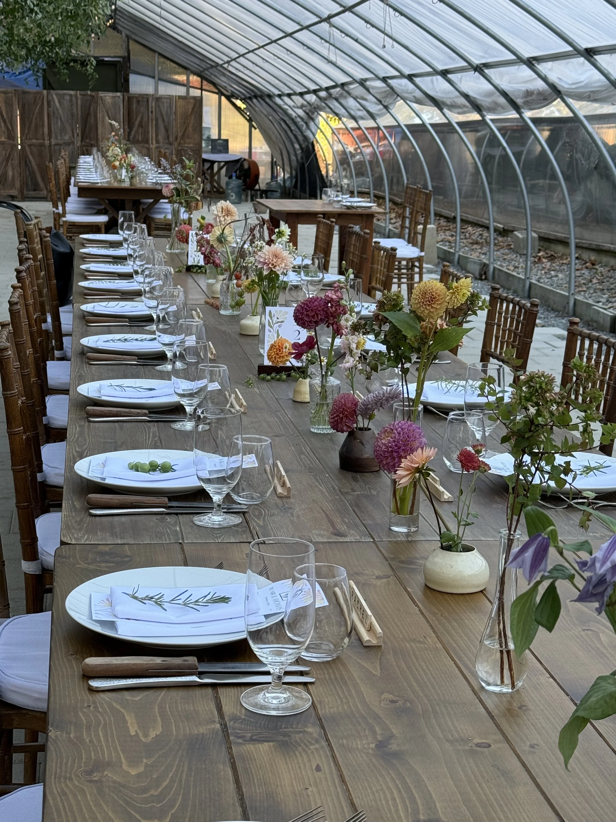 Long wooden table set for an outdoor event in a greenhouse, decorated with pink, purple, and yellow flowers in vases, with white plates, napkins, silverware, and glassware, and surrounded by wooden chairs.