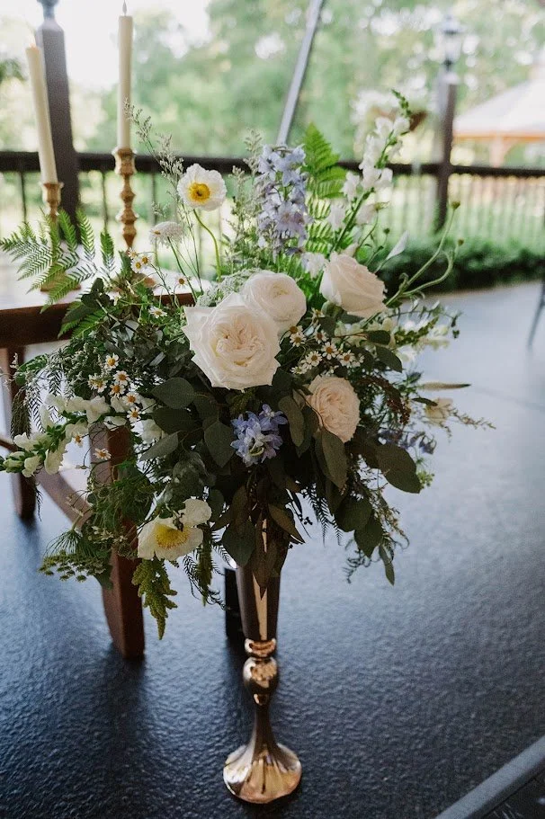 Flower arrangement with white roses, daisies, and greenery in a tall, elegant gold vase on a black table, with candles and a porch railing in the background.