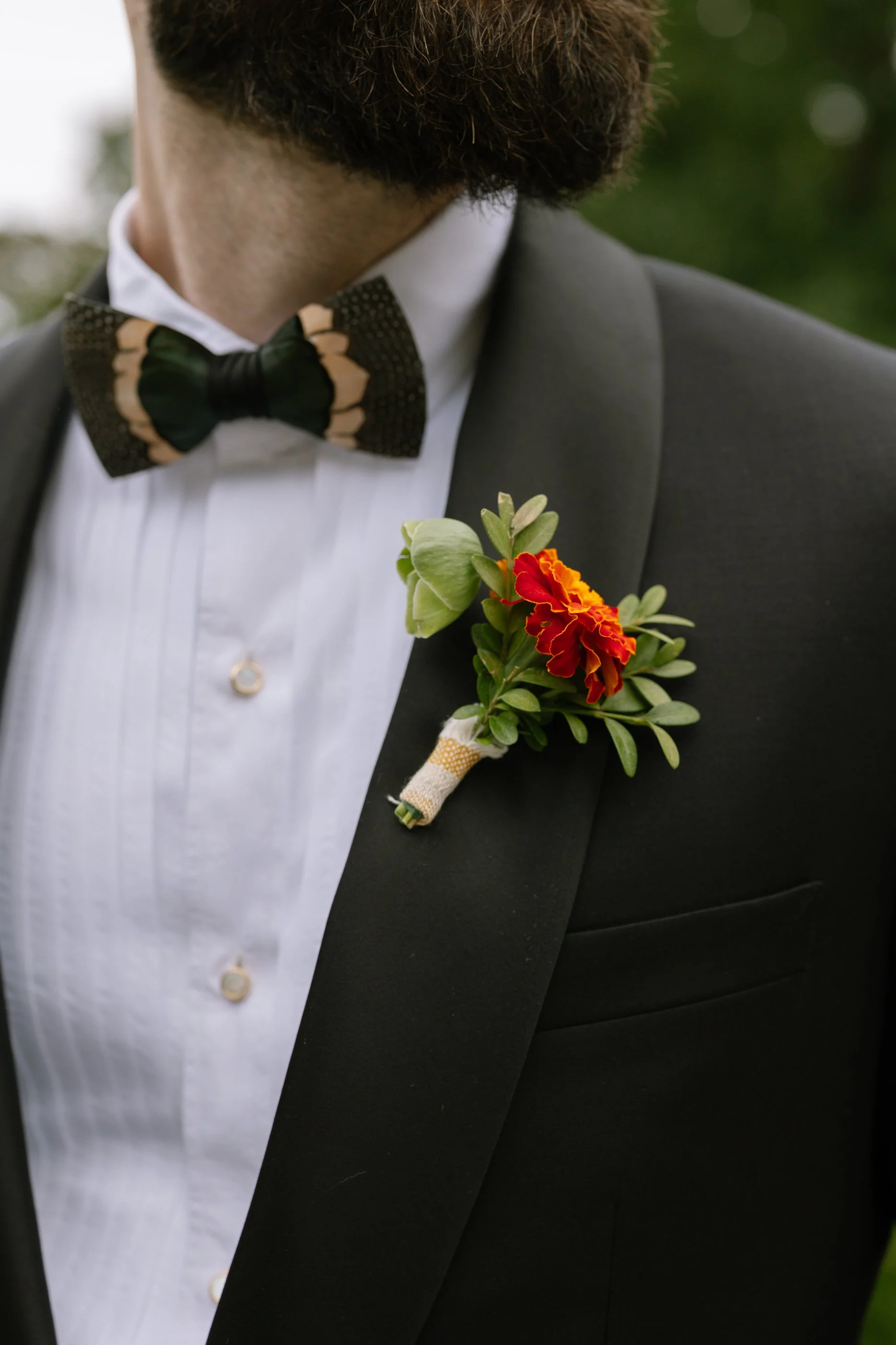 Close-up of a man's tuxedo with a boutonniere featuring a red and yellow flower, wearing a white dress shirt and a patterned black and beige bow tie.
