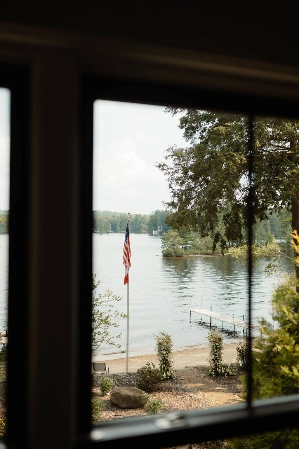 View of a lakeside with a flagpole flying the United States flag, a small dock extending into the water, and greenery, seen through a window frame.