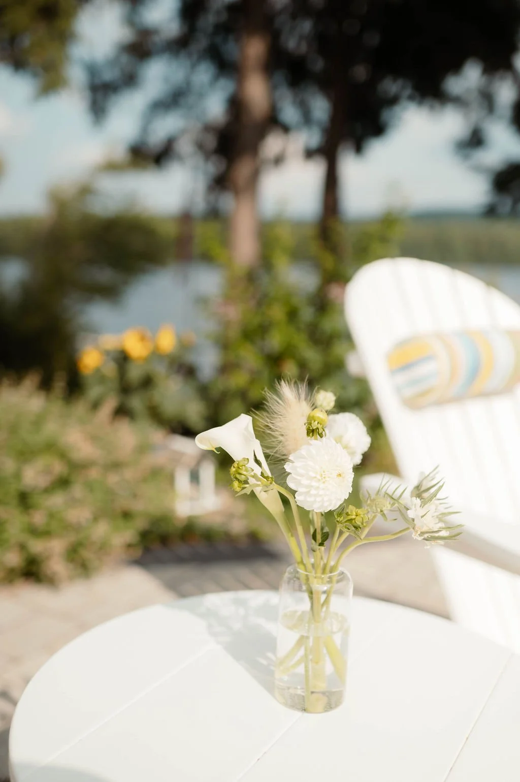 A small glass vase with white flowers and greenery on a white round table outdoors near a lake, with a white Adirondack chair in the background and trees.