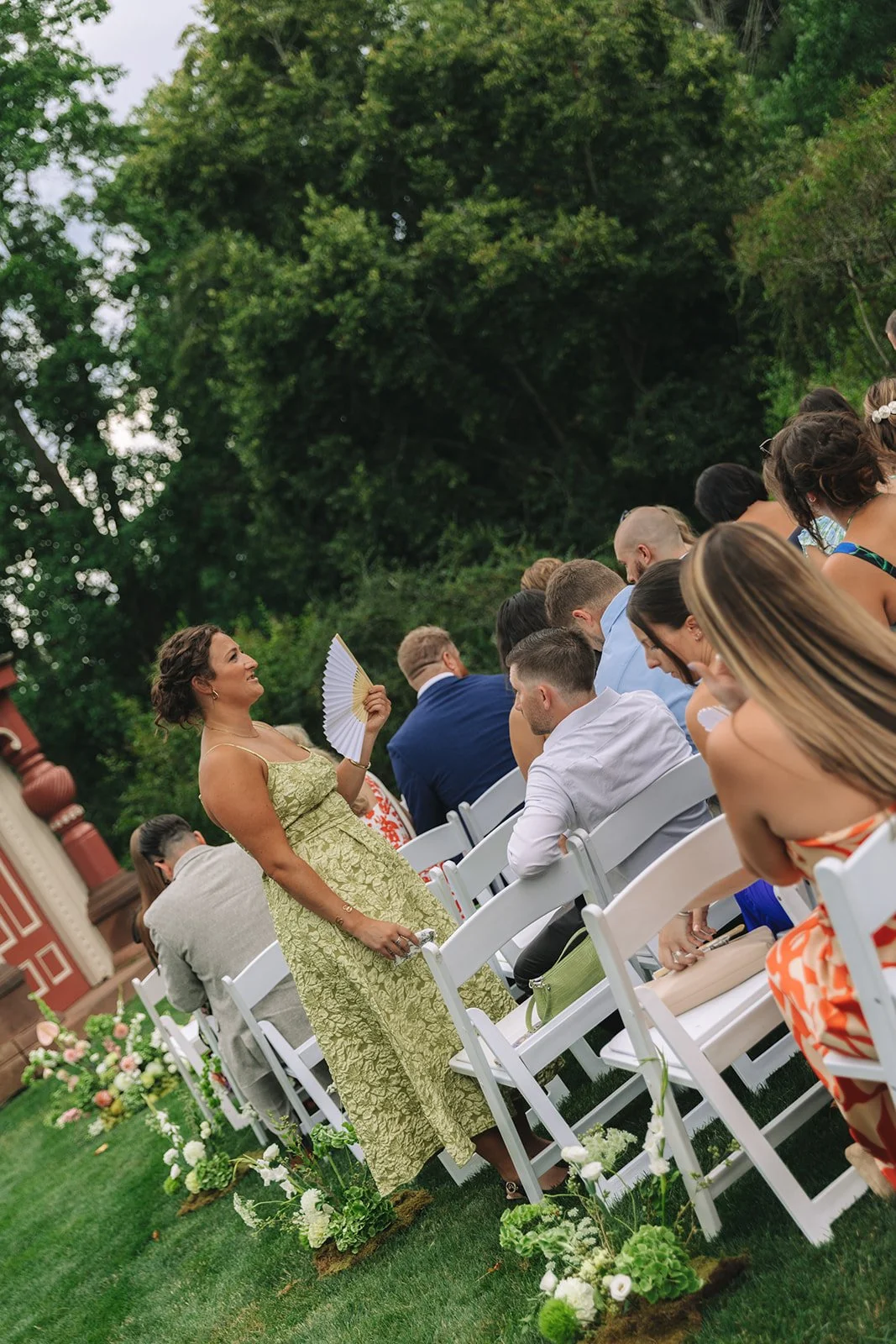 Outdoor wedding ceremony with seated guests, a woman in a light green dress holding a fan, and lush green trees in the background.