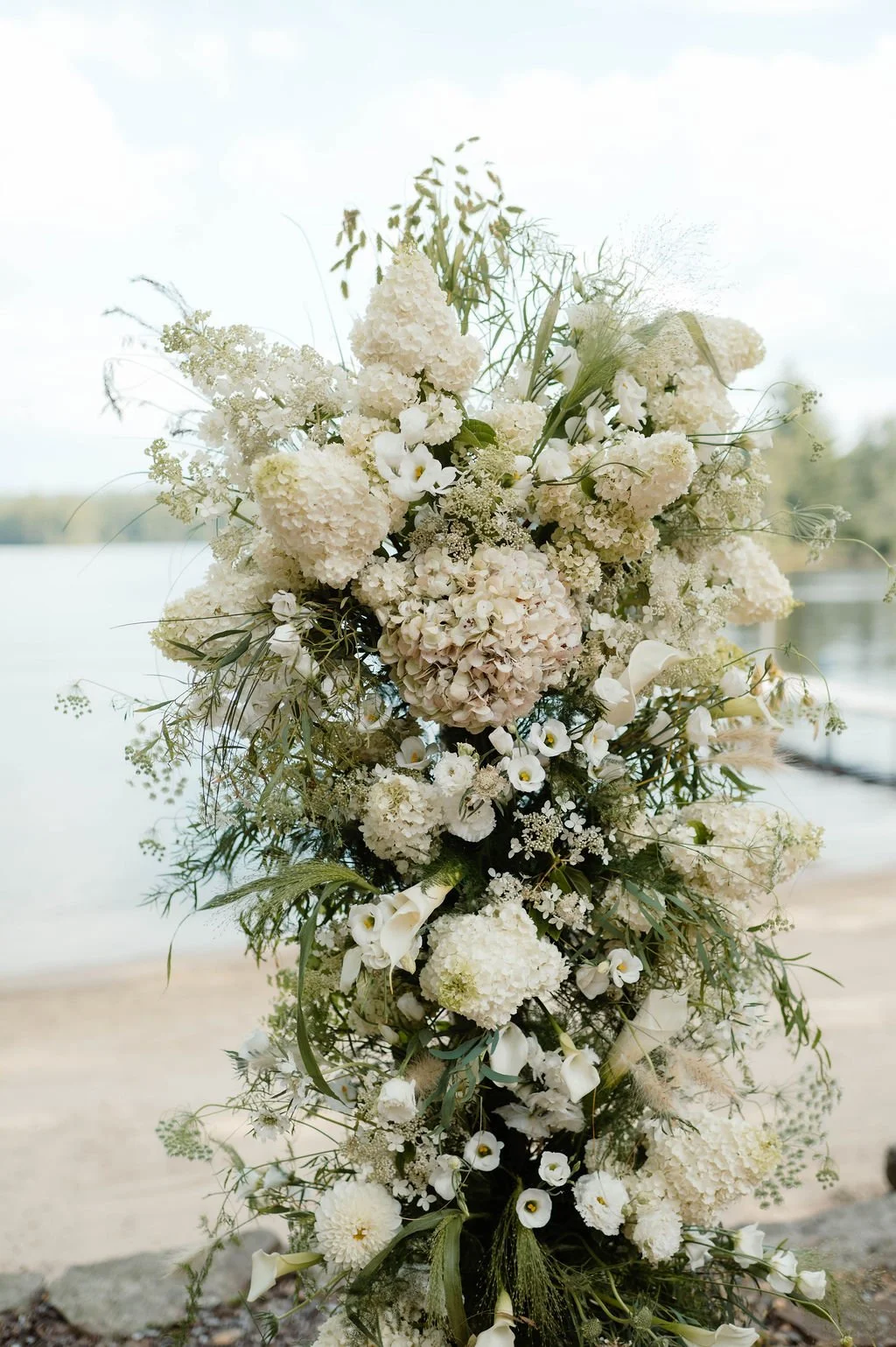 A large white floral arrangement with various flowers and greenery, set outdoors near a lake.