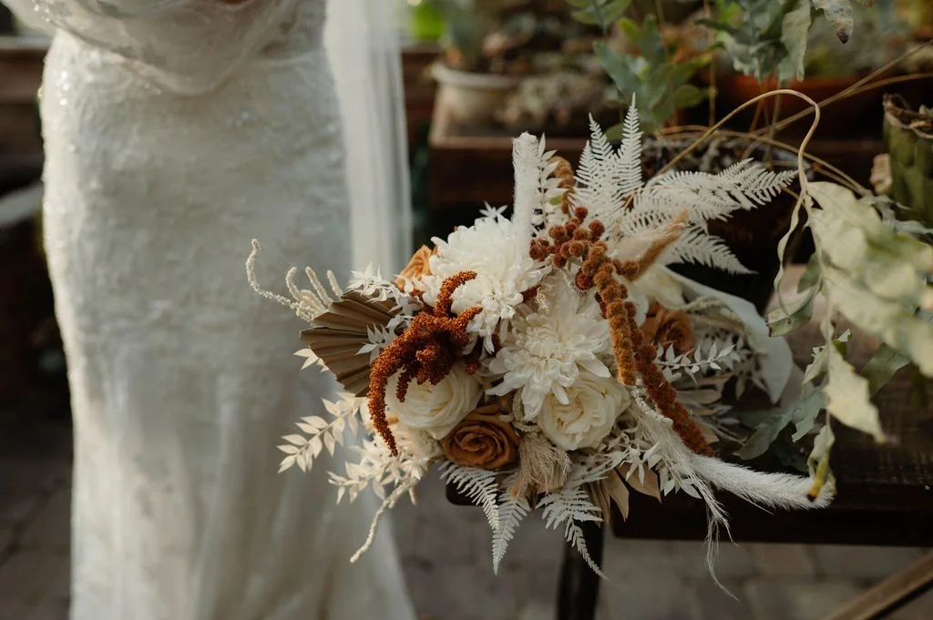A dried flower bouquet with white, cream, and brown flowers and foliage, placed on a rustic wooden table next to a woman in a lace dress.