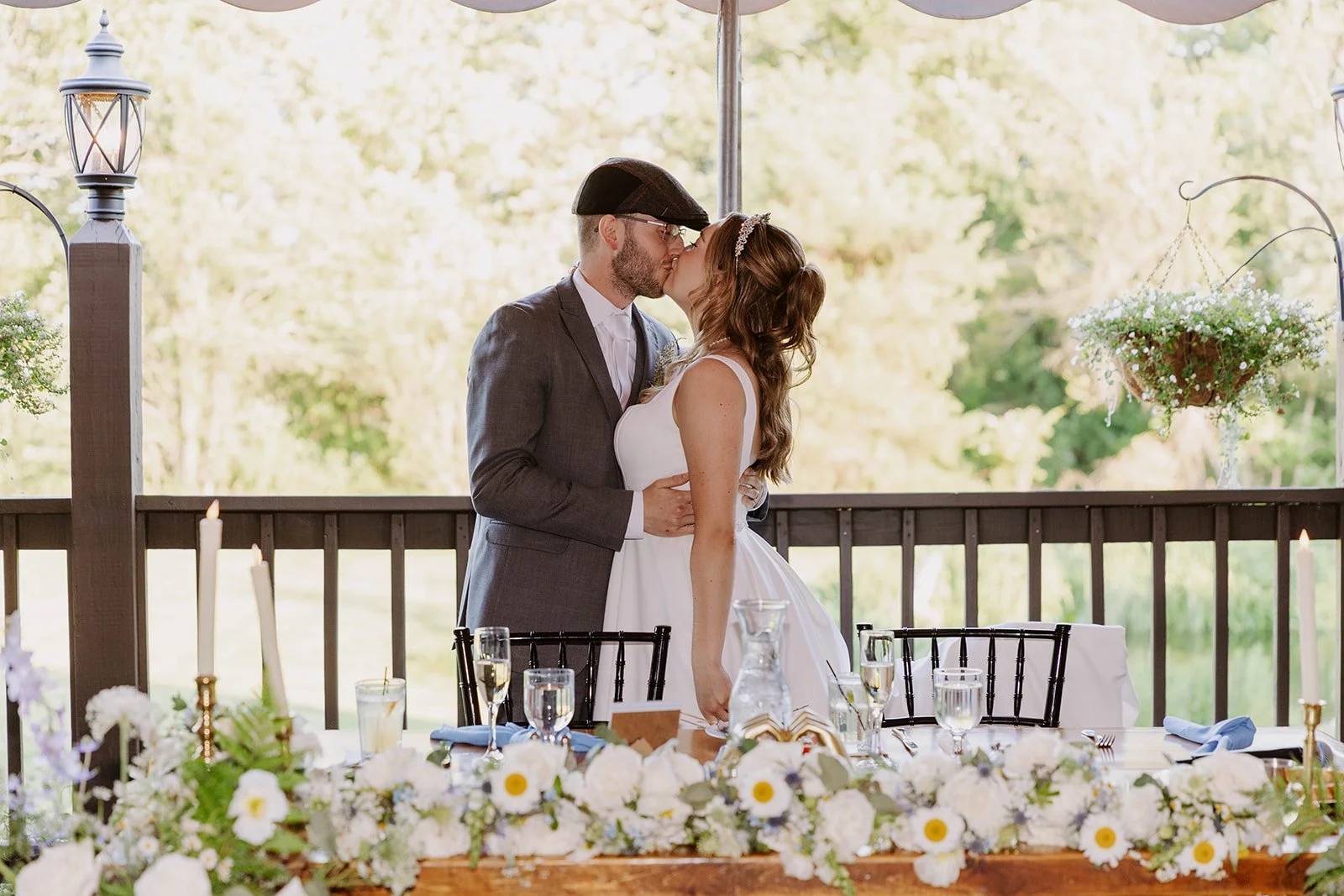 A bride and groom sharing a kiss at their wedding reception on a decorated outdoor wooden deck, with a table adorned with white flowers and candles in the foreground.
