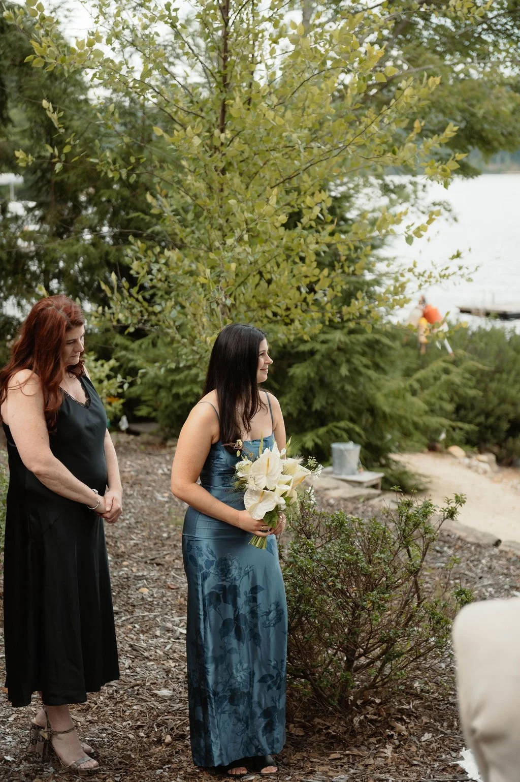 A woman in a blue floral dress holding a bouquet of white lilies stands outdoors during a ceremony, with another woman in a black dress beside her, near trees and a body of water.