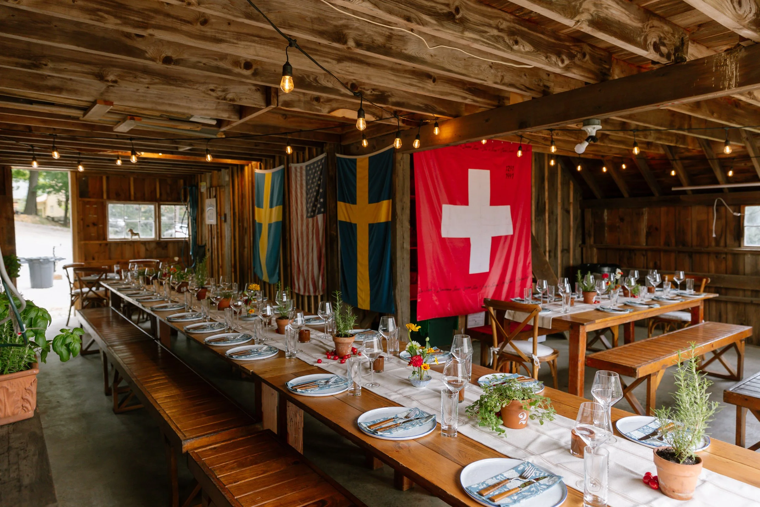 Long wooden dining tables set for a meal with plates, glasses, and silverware, decorated with potted plants and flowers, inside a rustic wooden structure with string lights and flags from various countries hanging on the wall.