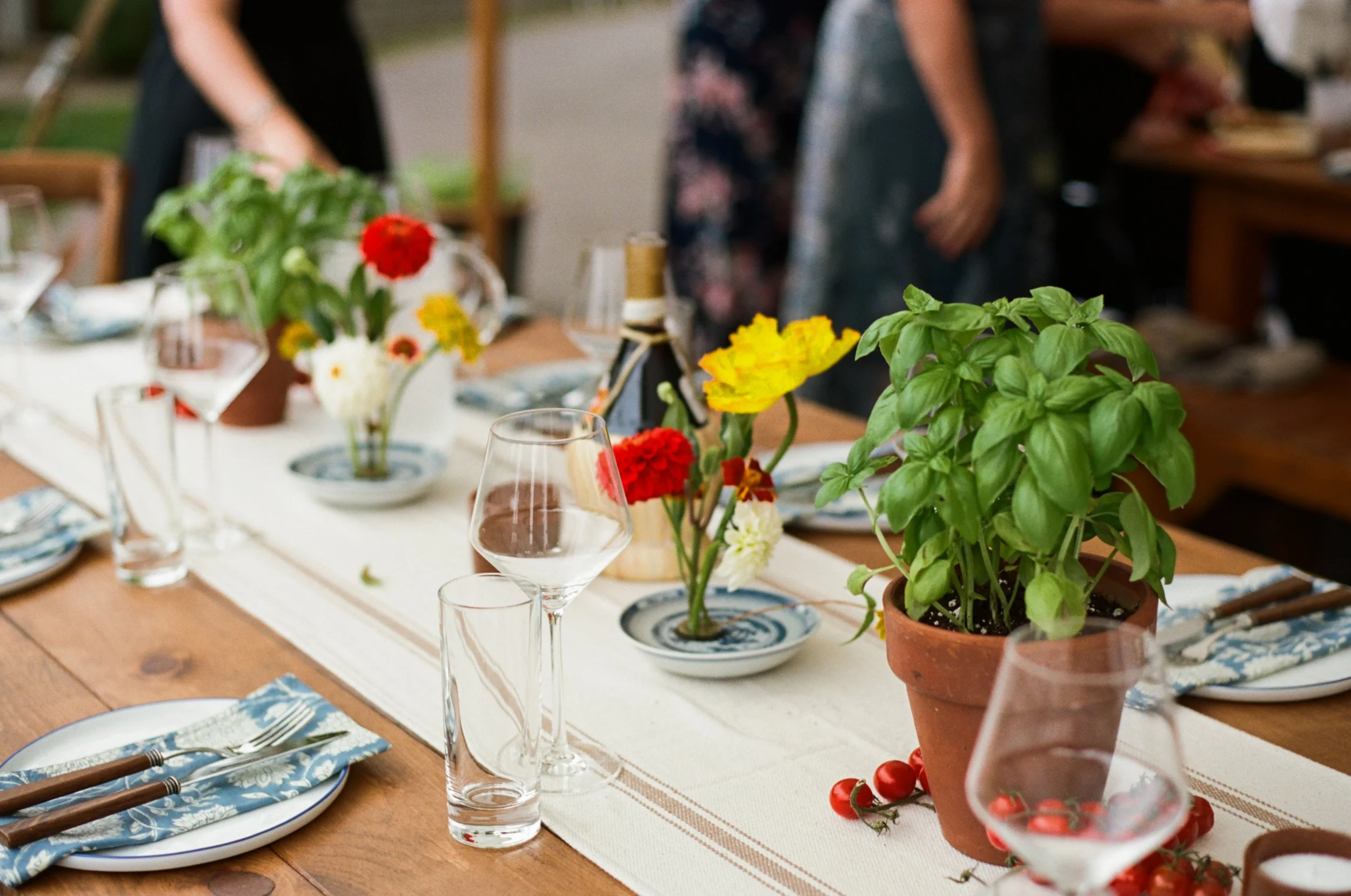 Table set for a meal with colorful flowers and potted herbs, including basil, on a white table runner. Glassware and plates with blue and white patterns are arranged, with people gathering in the background.