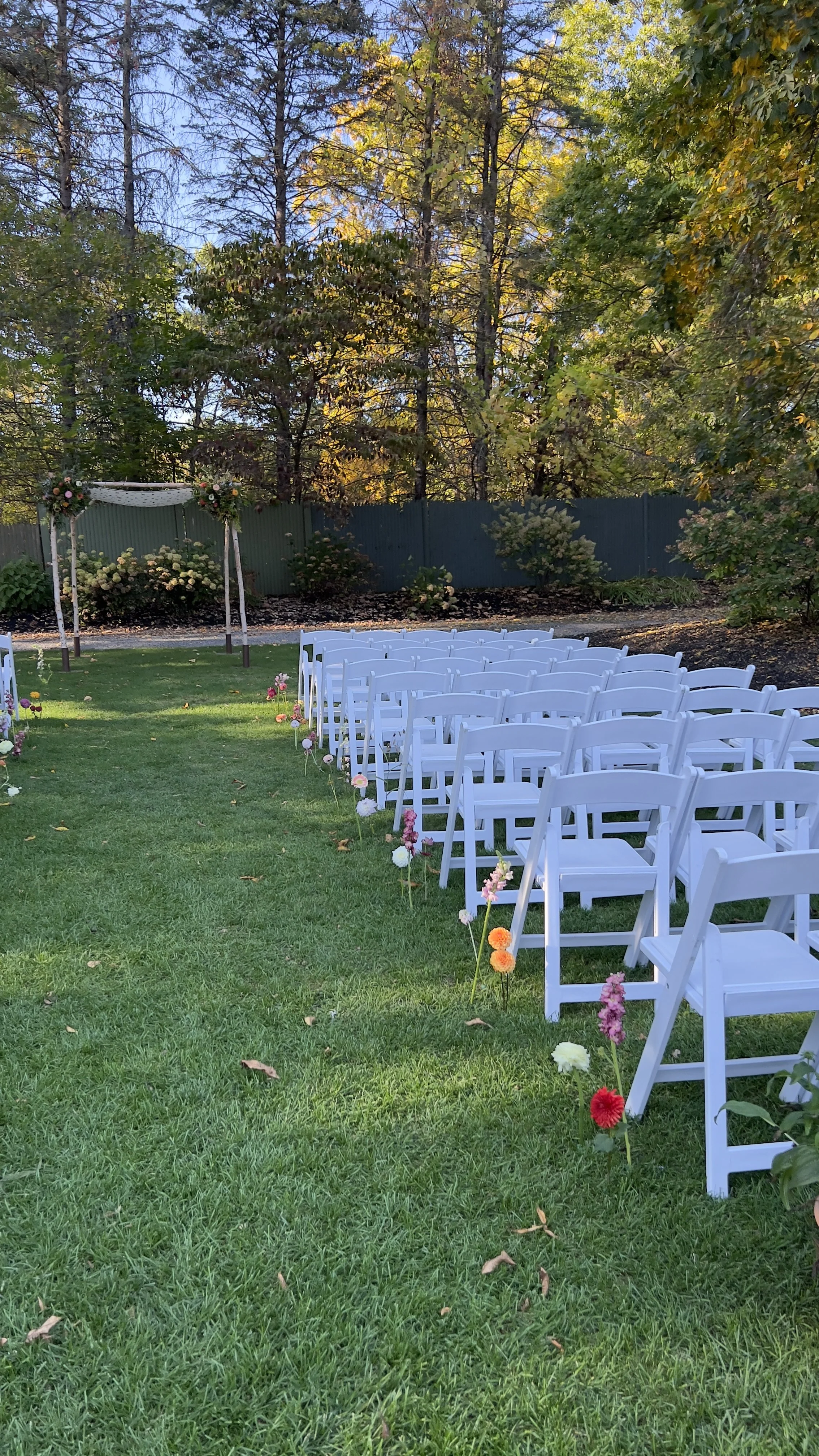 Outdoor wedding setup with white chairs arranged in rows, decorated with colorful flowers, and a floral arch in the background, set in a grassy area surrounded by trees with autumn foliage.