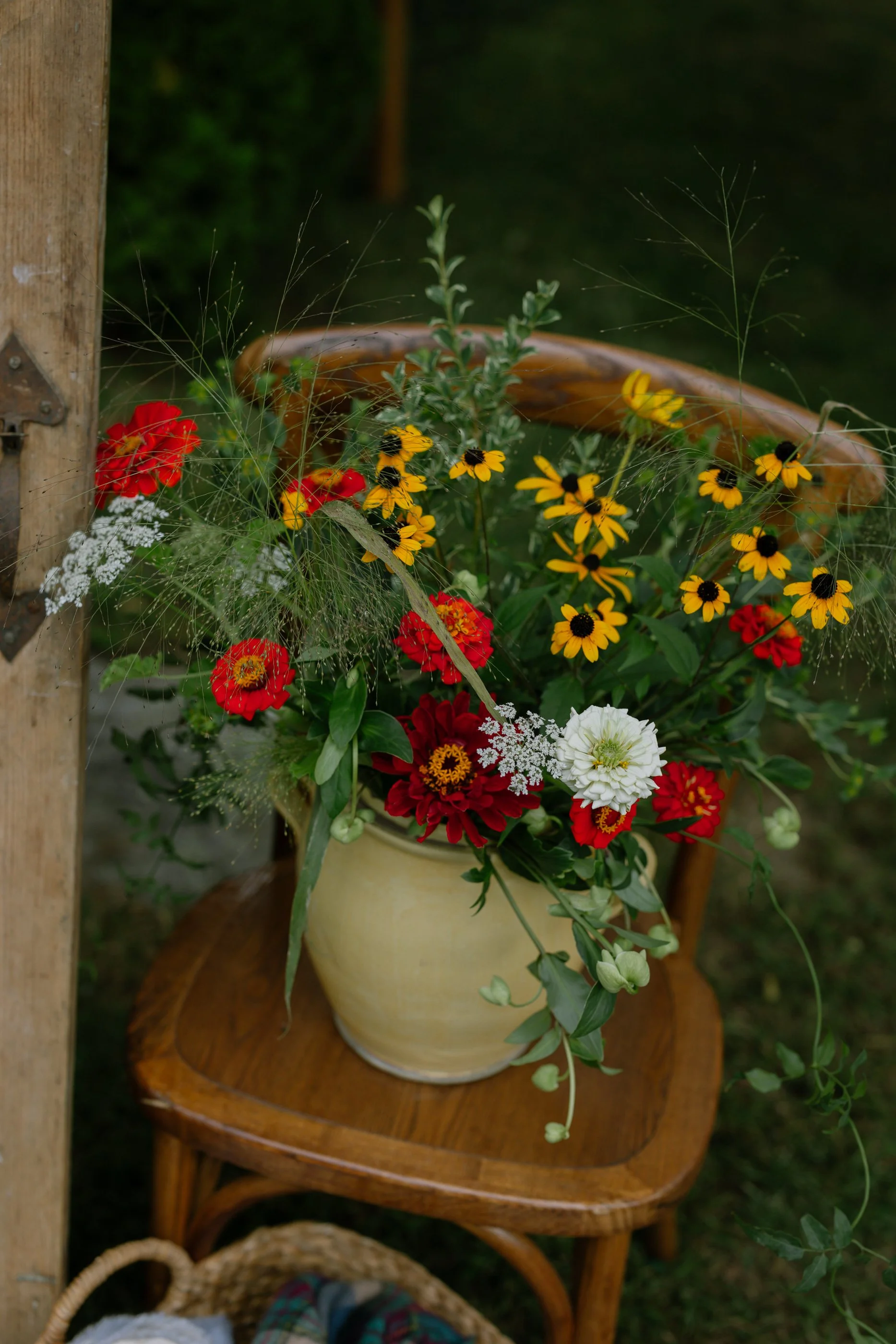 A bouquet of red, white, and yellow flowers in a beige ceramic vase on a wooden chair outdoors.