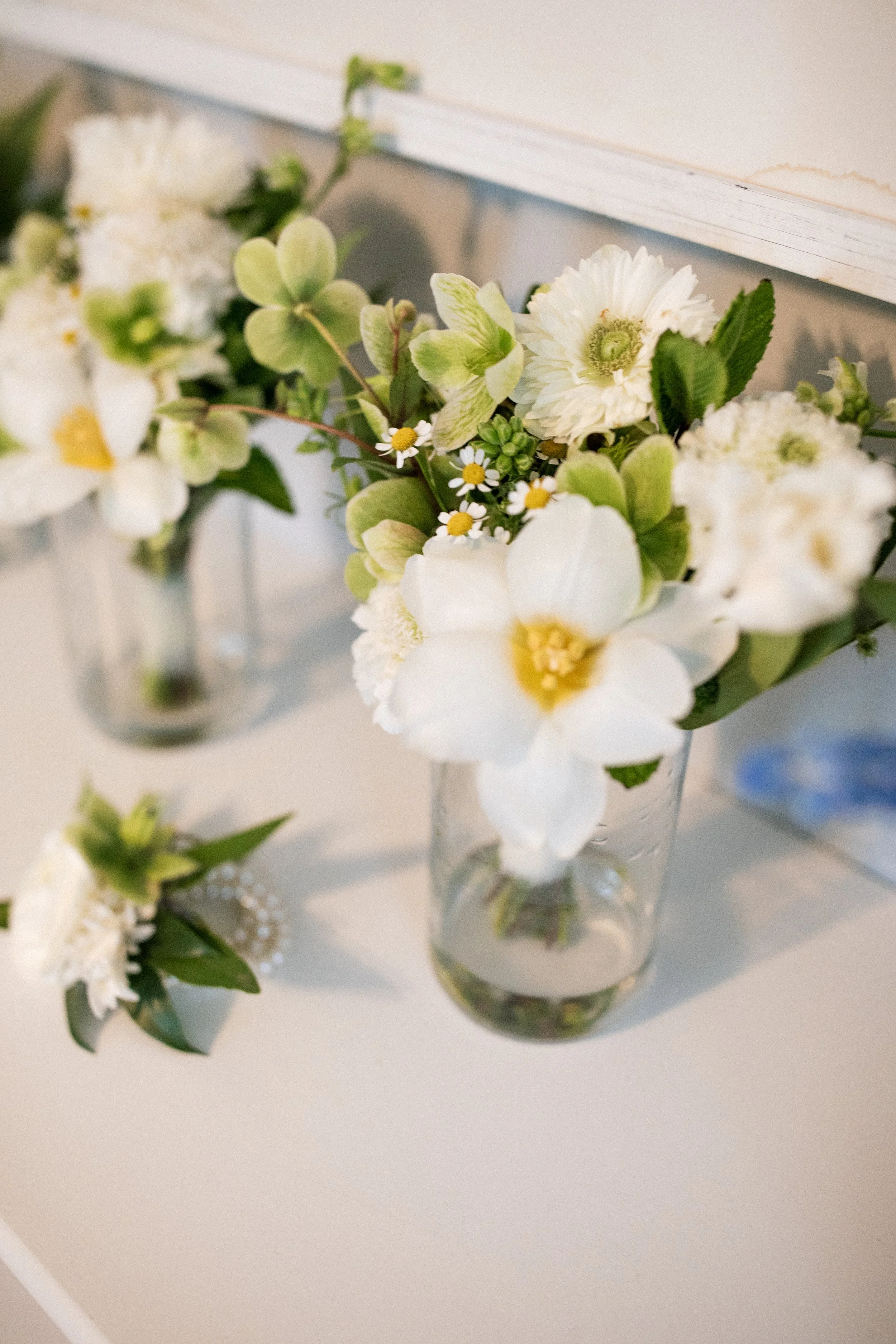 A clear glass vase with white flowers and green leaves on a white table, with a similar arrangement in the background.