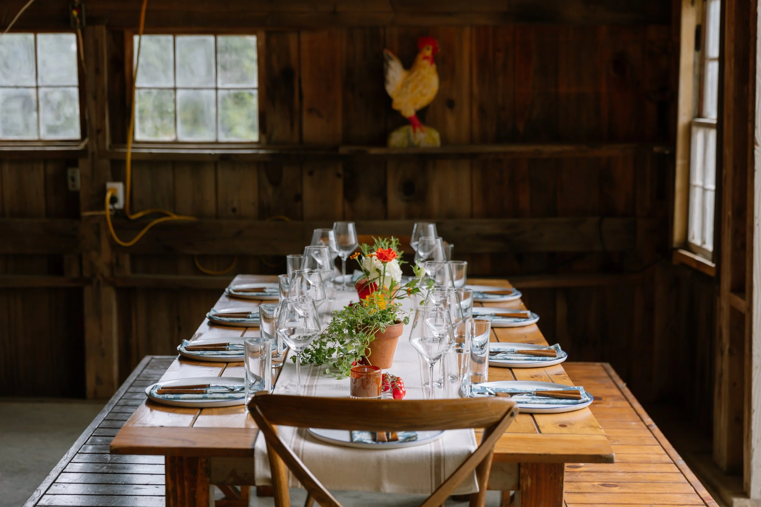 A rustic wooden dining table set for a meal with glassware, plates, and cutlery, decorated with potted plants and small flower arrangements, inside a wooden structure with windows and a decorative chicken wall hanging.