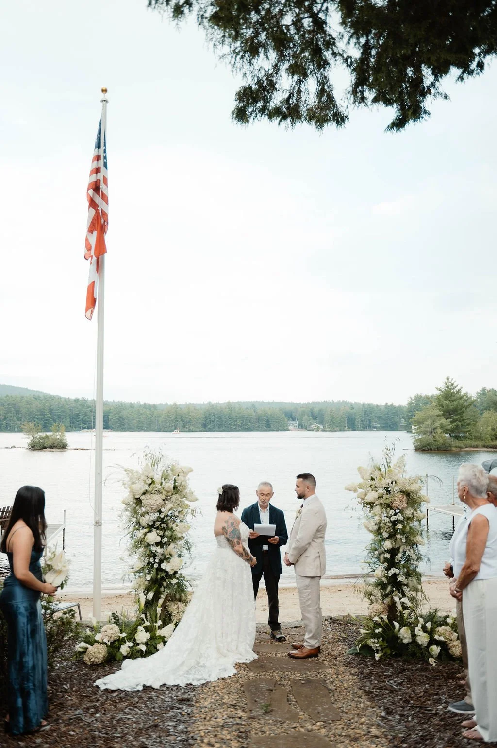 A wedding ceremony taking place outdoors by a lake, with officiant, bride, groom, bridesmaid, and guests, all dressed formally, surrounded by floral arrangements and an American flag on a flagpole.