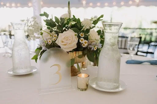 Table setting with a floral centerpiece of white roses and daisies, two glass vases, and a small candle holder, set outdoors or in a bright indoor space.