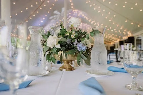 Table centerpiece with white flowers and greenery in a gold vase, flanked by two glass water pitchers, with blue napkins on a white tablecloth, under string lights.