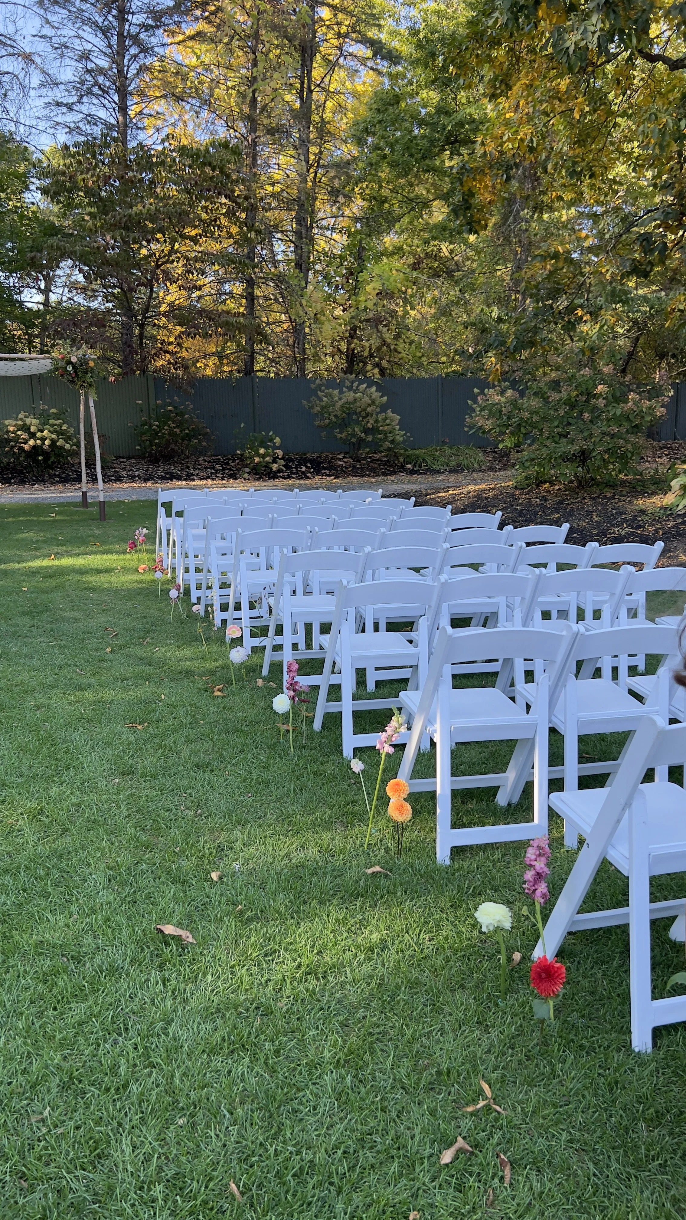 Rows of white chairs arranged outdoors for a wedding ceremony, decorated with colorful flowers along the edge of the grass, in a garden setting with trees and a fence in the background.