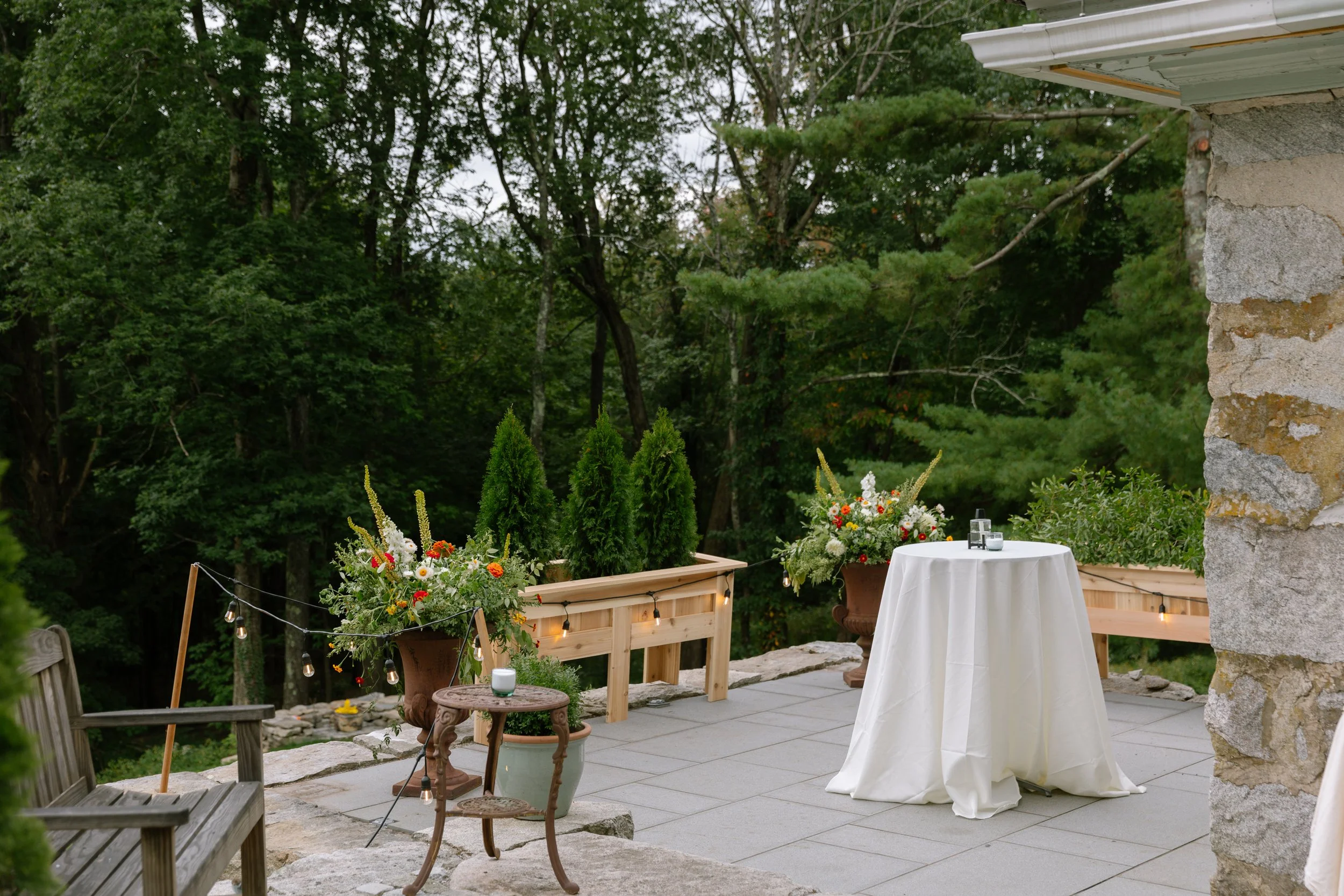 Outdoor patio decorated with potted flowers, a round table with a white tablecloth, and string lights, surrounded by lush green trees and stone features.