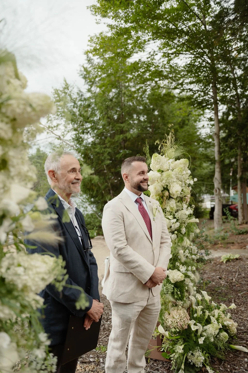 A groom in a beige suit with a red tie standing outdoors near a large floral arrangement during a wedding ceremony, smiling, with an officiant or guest in a dark jacket standing next to him.